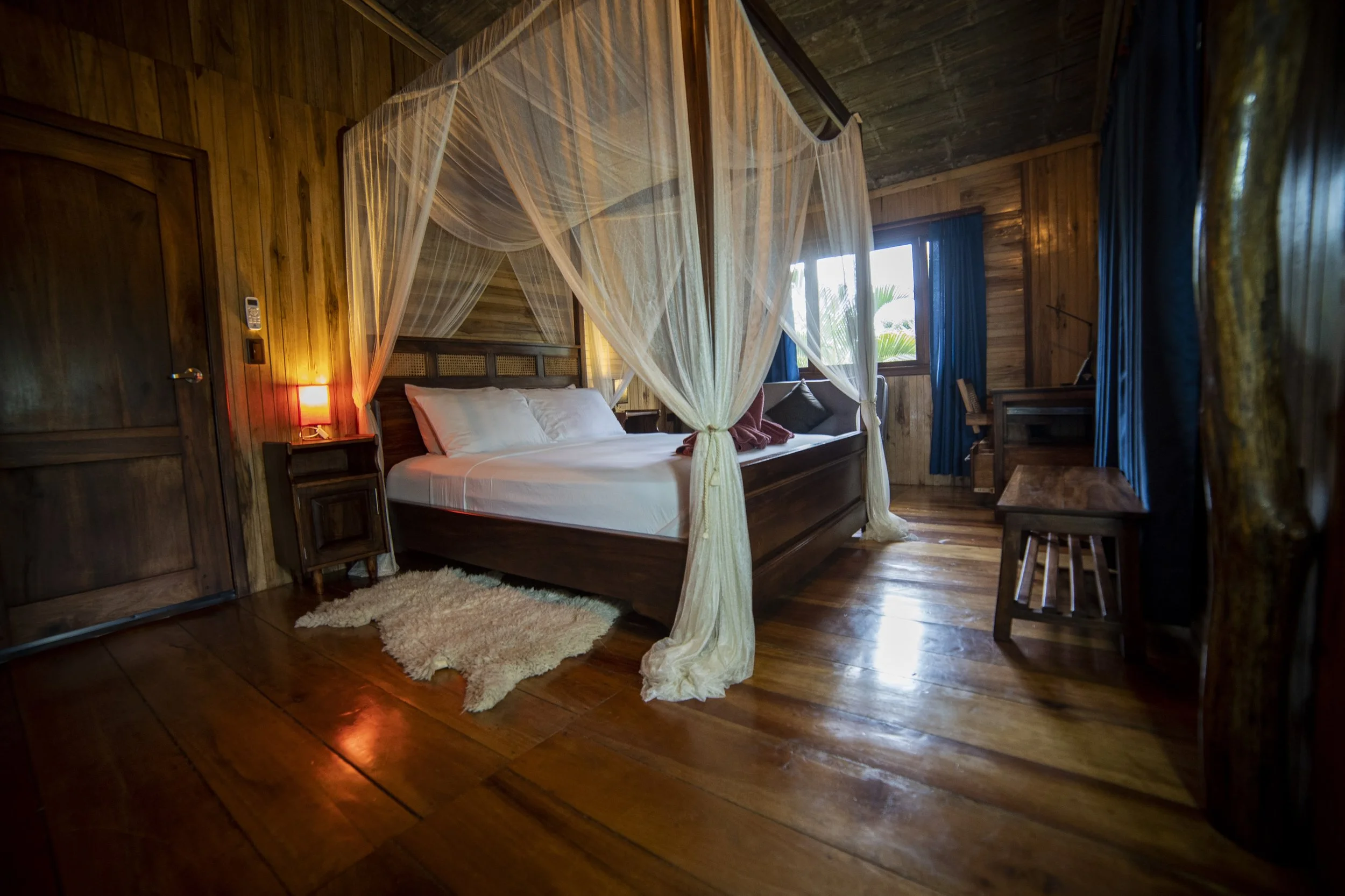 Habitación con cama de madera y cortinas de mosquitero, paredes de madera, piso de madera, una lámpara con luz cálida, ventanas con cortinas azules, y un área de trabajo con escritorio.