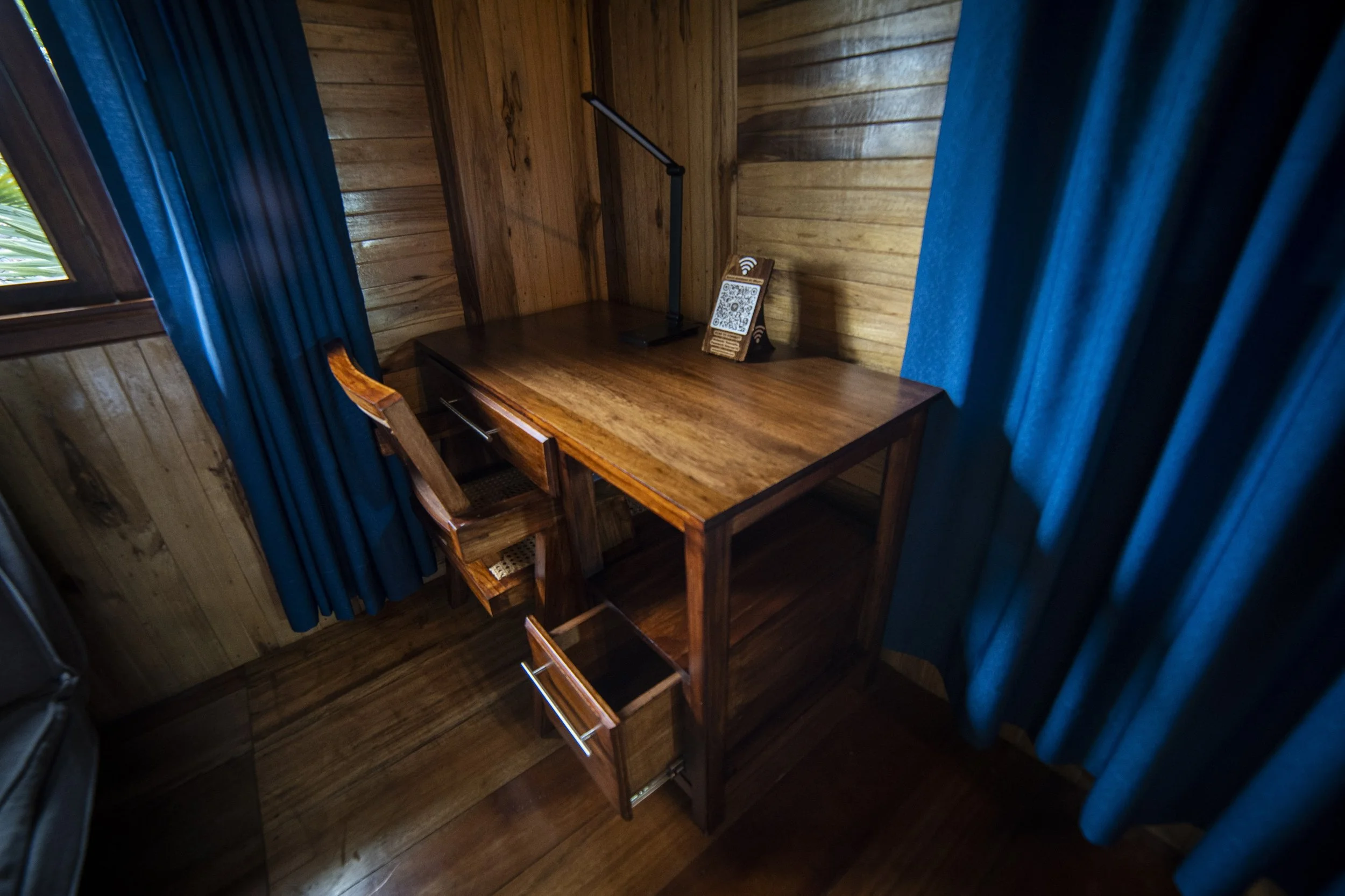 escritorio de madera con una lámpara y un código QR, rodeado de cortinas azules en una habitación con paredes y piso de madera