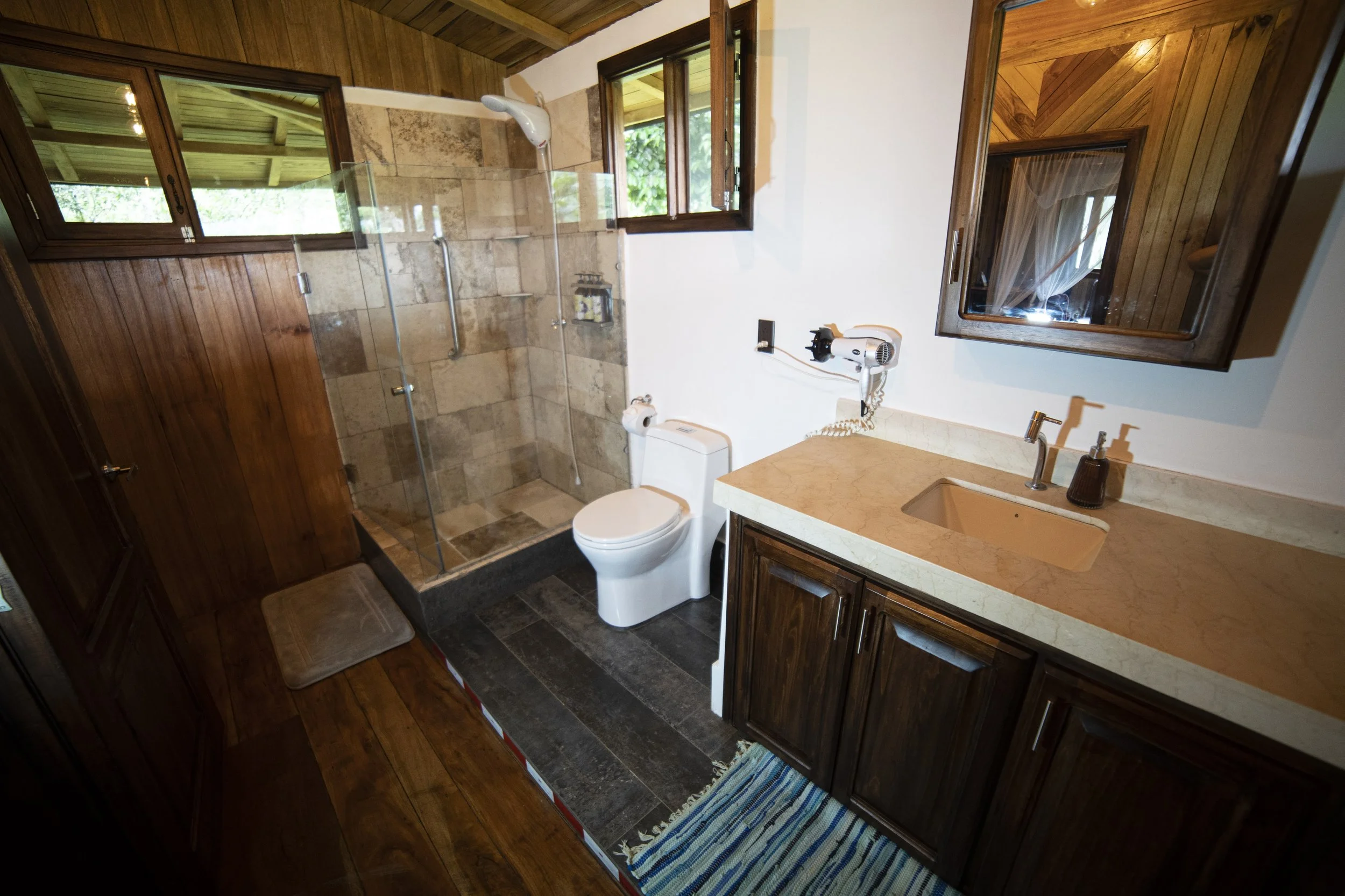 Un baño con paredes de madera y piso de piedra negra. Incluye un lavabo con encimera color durazno, armario de madera oscuro, espejo de madera, inodoro y ducha con puertas de vidrio. Hay ventanas abiertas con vista a árboles verdes.