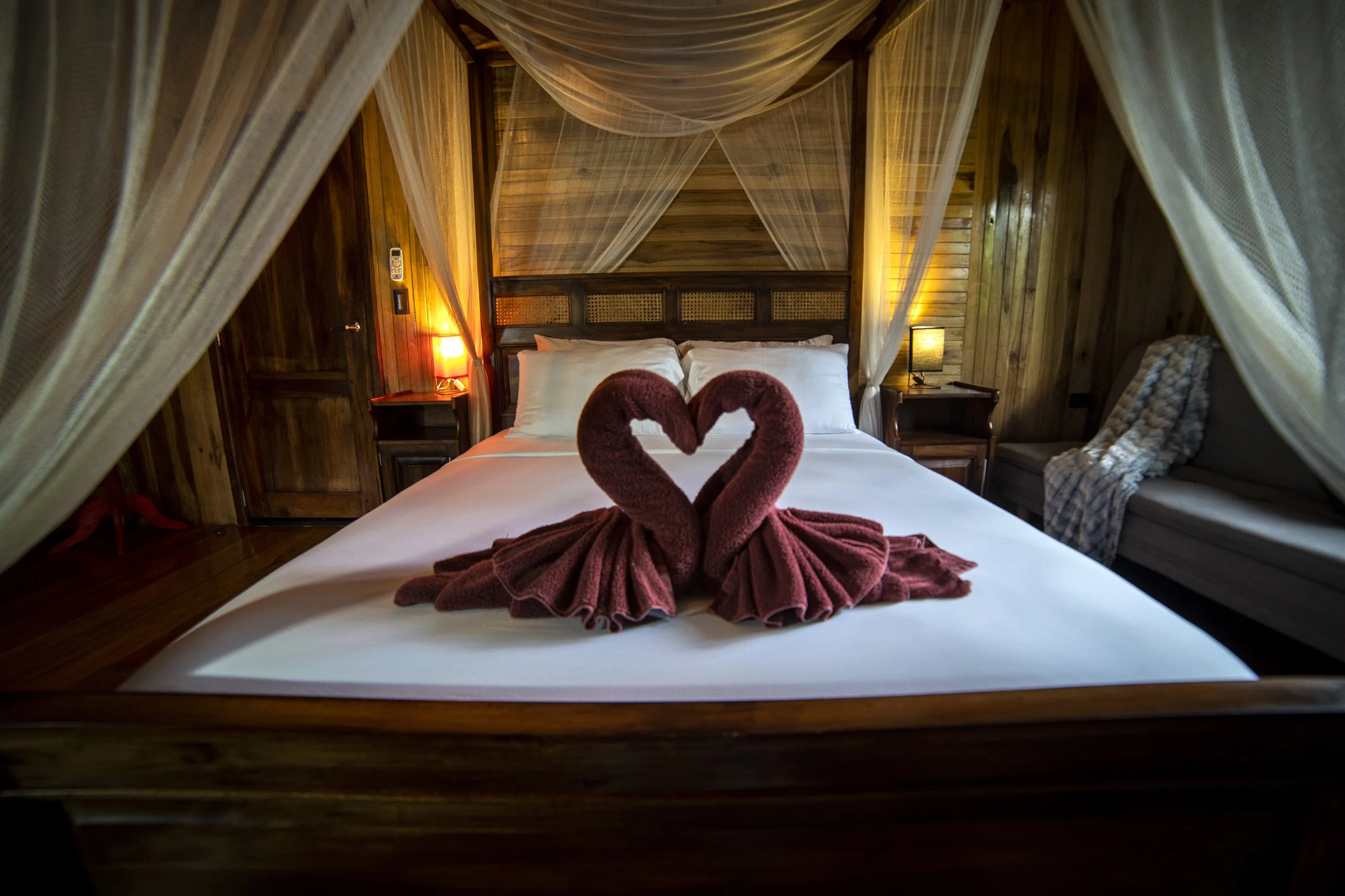 Cama de madera con sábanas blancas, decorada con toallas en forma de corazones de color rosa que forman un par de cisnes, en una habitación con paredes de madera y cortinas ligeras.