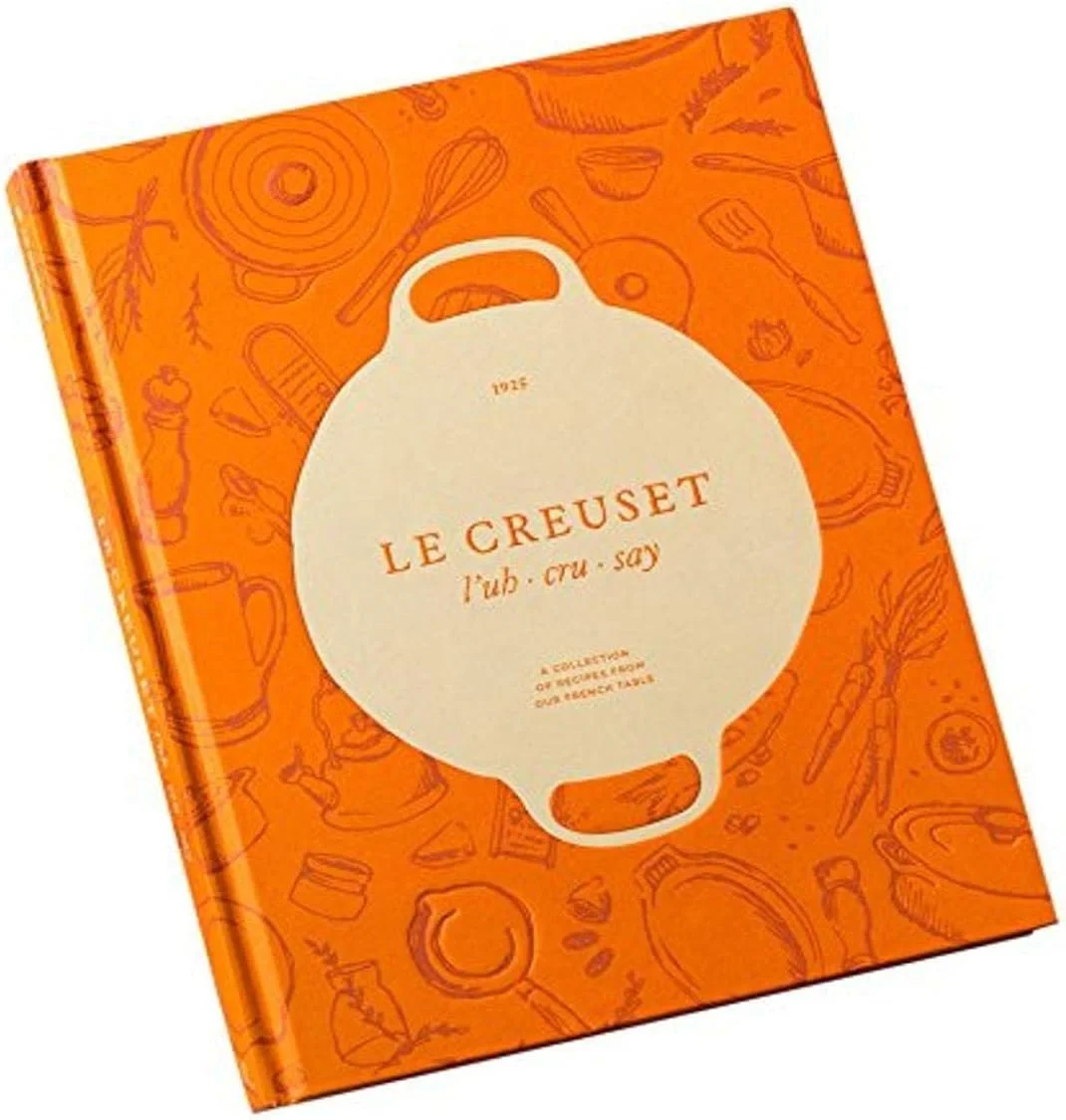 Libro de cocina de color naranja con ilustraciones de utensilios y ingredientes, y un círculo central que dice 'Le Creuset'.