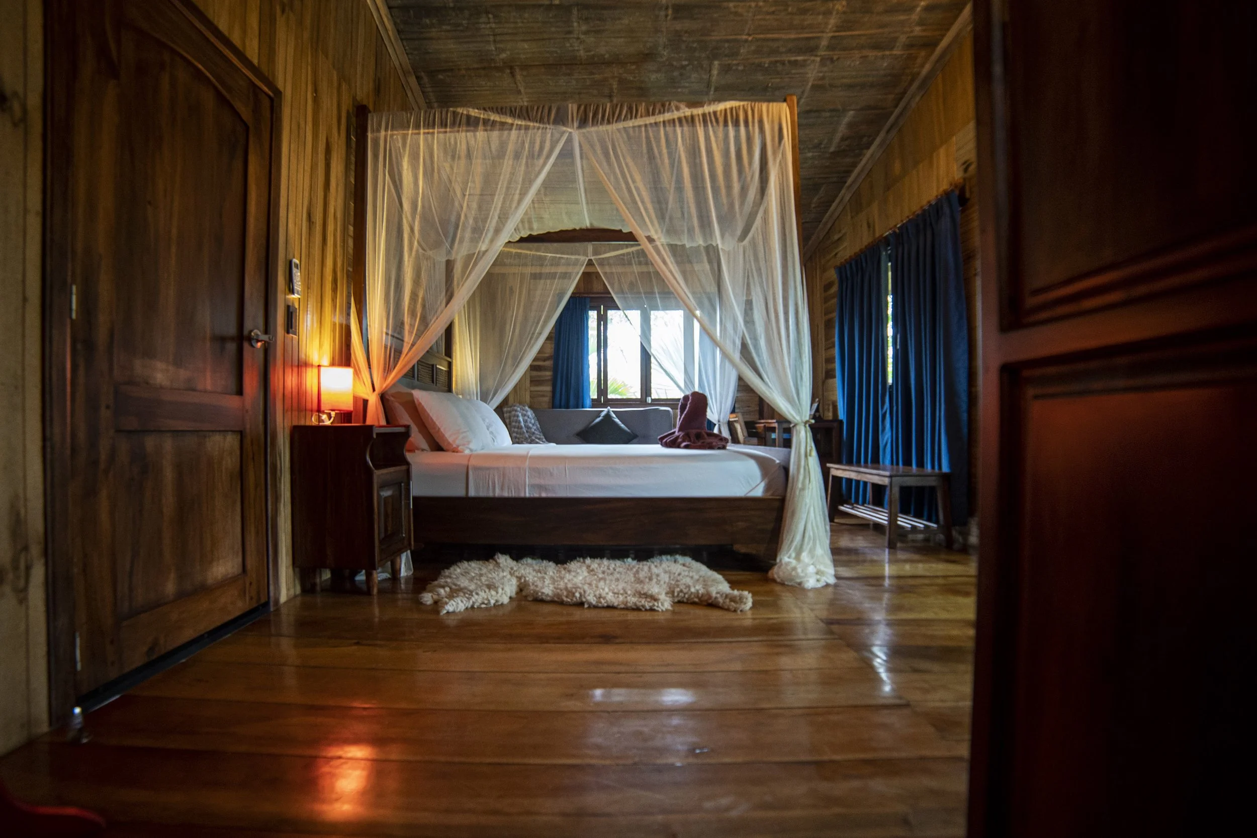 Habitación de hotel con paredes y suelo de madera, cama con dosel y cortinas blancas, almohadas blancas y decoración acogedora con luz tenue, cortinas azules y una ventana al fondo.