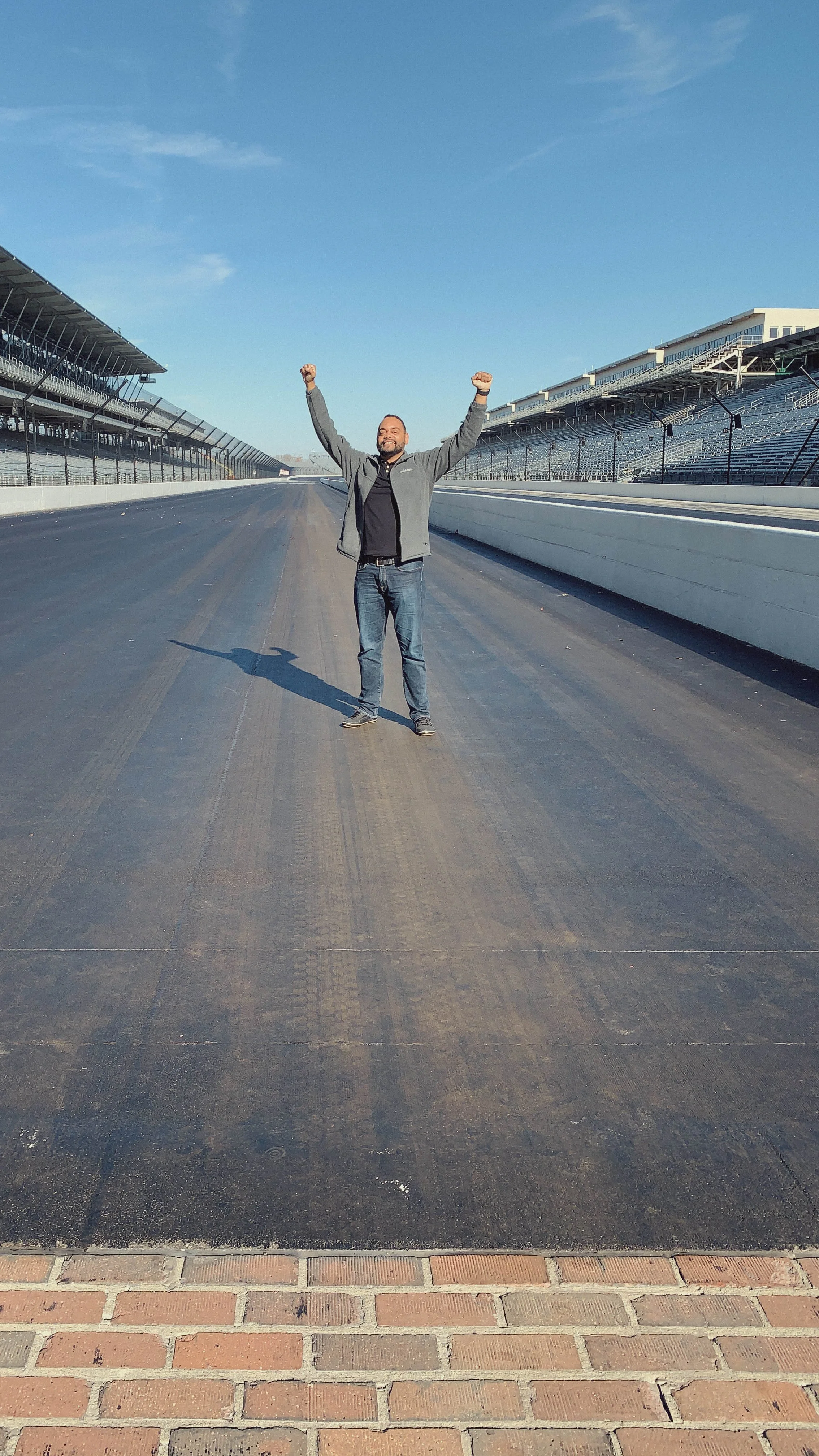 A man standing in the middle of a racetrack with arms raised in victory, sunny day, empty grandstands on either side.
