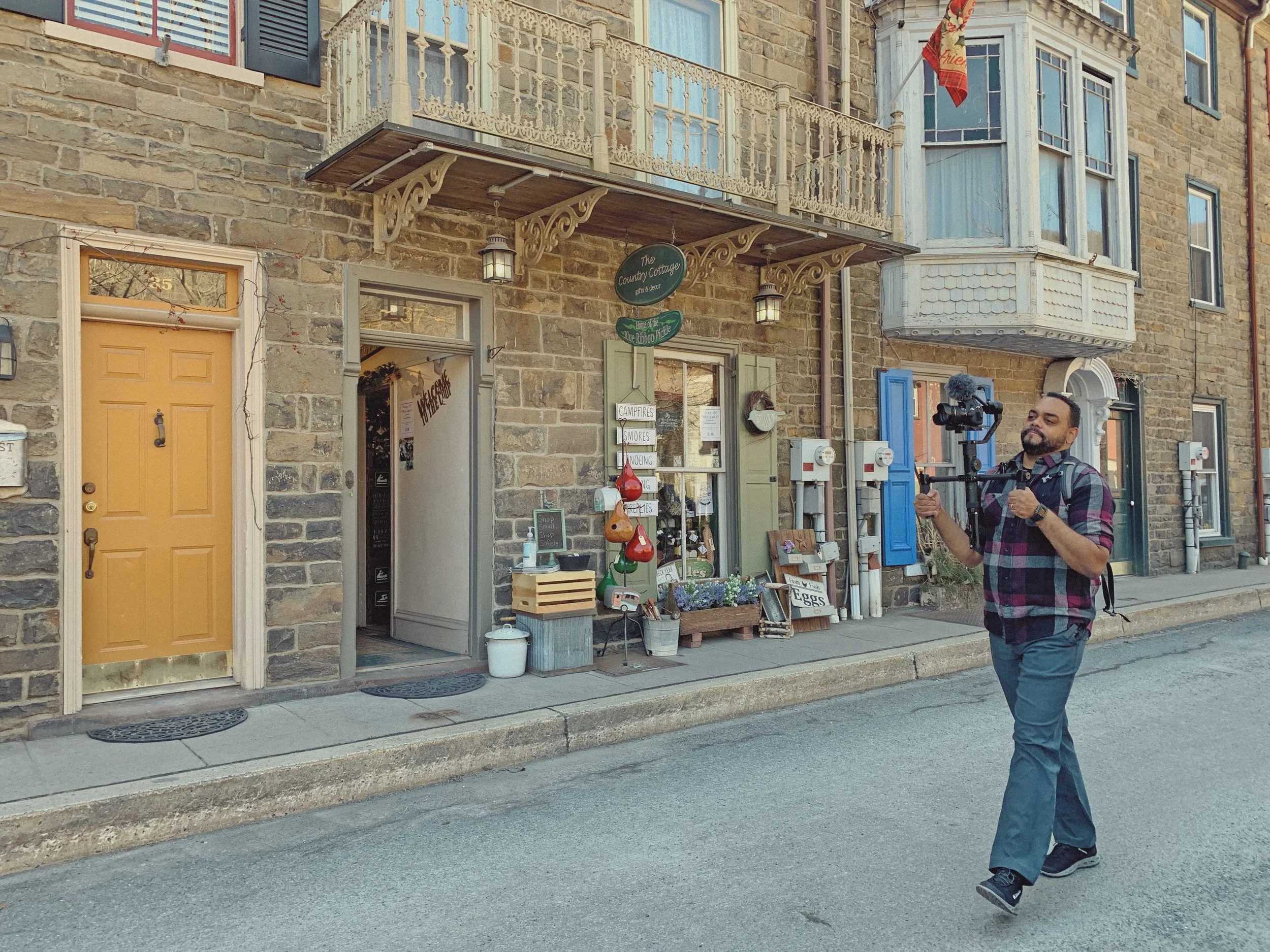 A man holding a camera with a stabilizer is walking past a storefront called The Country Cottage, which sells gifts and decor. The building has a stone facade, a yellow door, and a small outdoor display of plants and household items. The storefront h