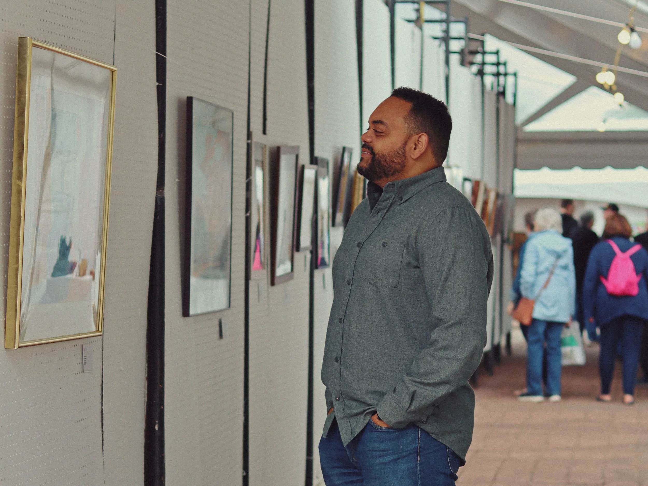 A man in a gray shirt and jeans looking at framed pictures or artwork on a wall at an art gallery or exhibition.