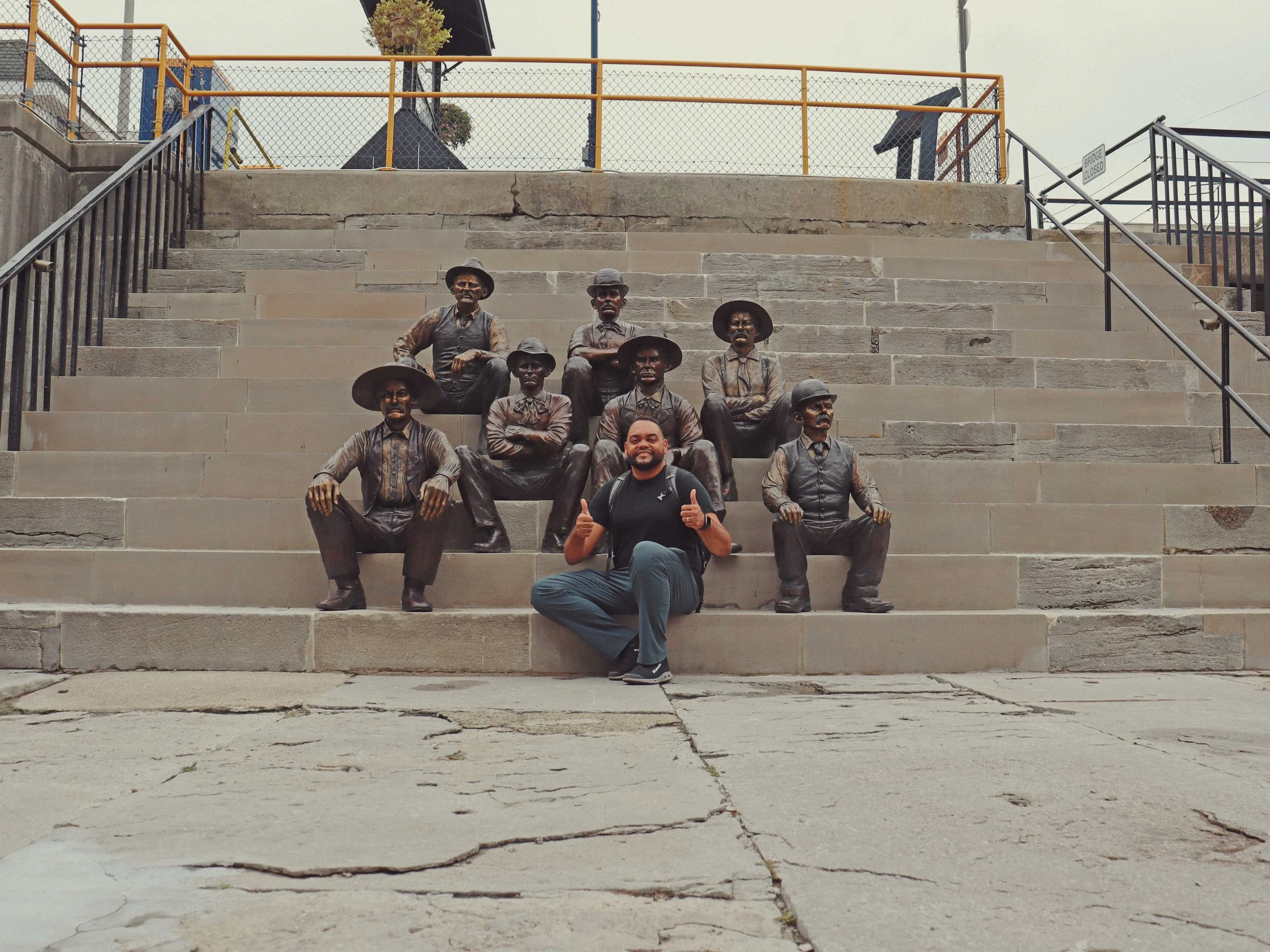 A man sitting on stairs in front of bronze statues of historical figures. The statues depict men in period clothing with hats, arranged in a group. The stairs are cracked and worn, with handrails on each side. Background features a chain-link fence a