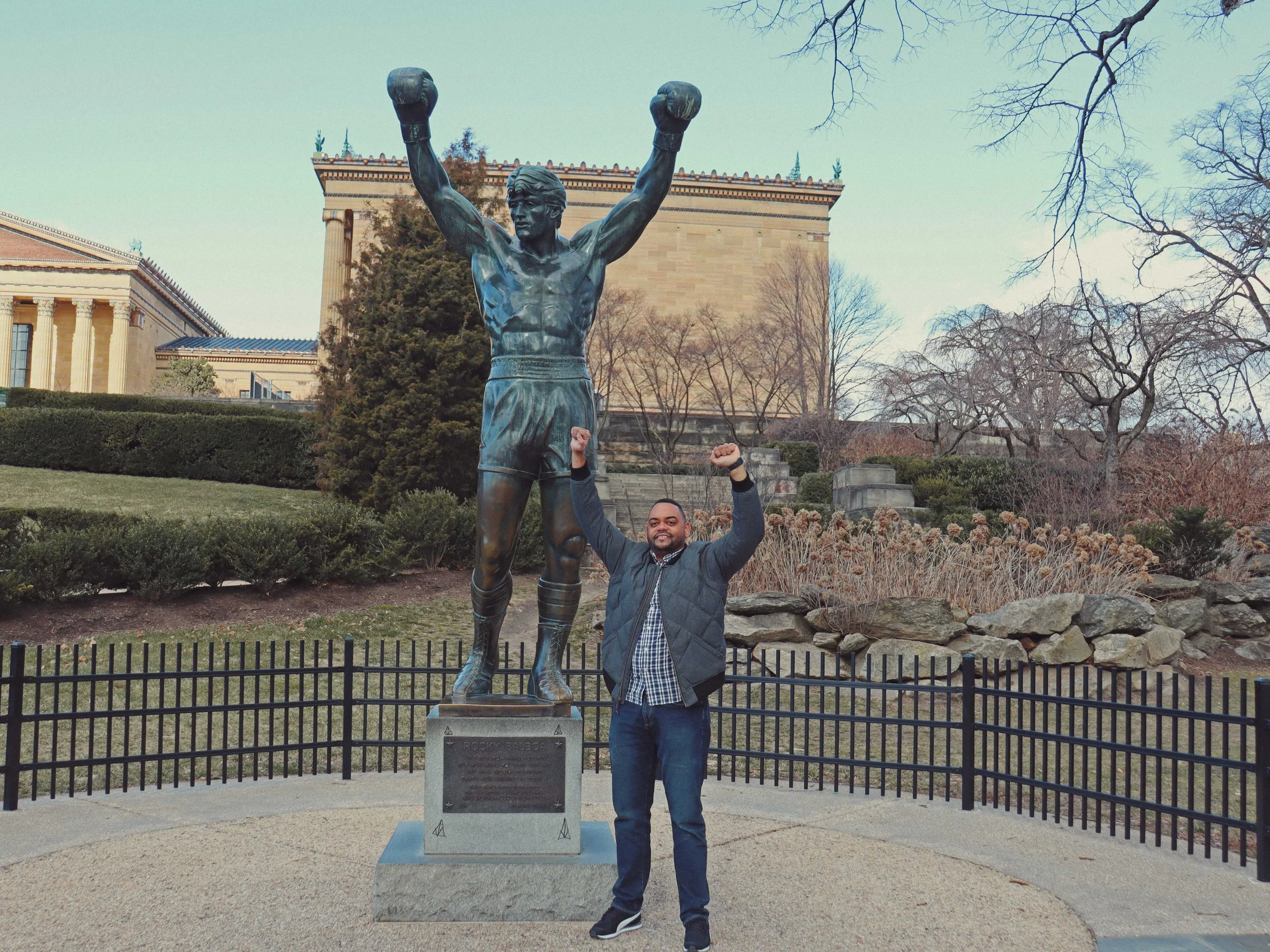 A man standing next to the Rocky Balboa statue at the Philadelphia Museum of Art, raising his arm in a boxing pose.