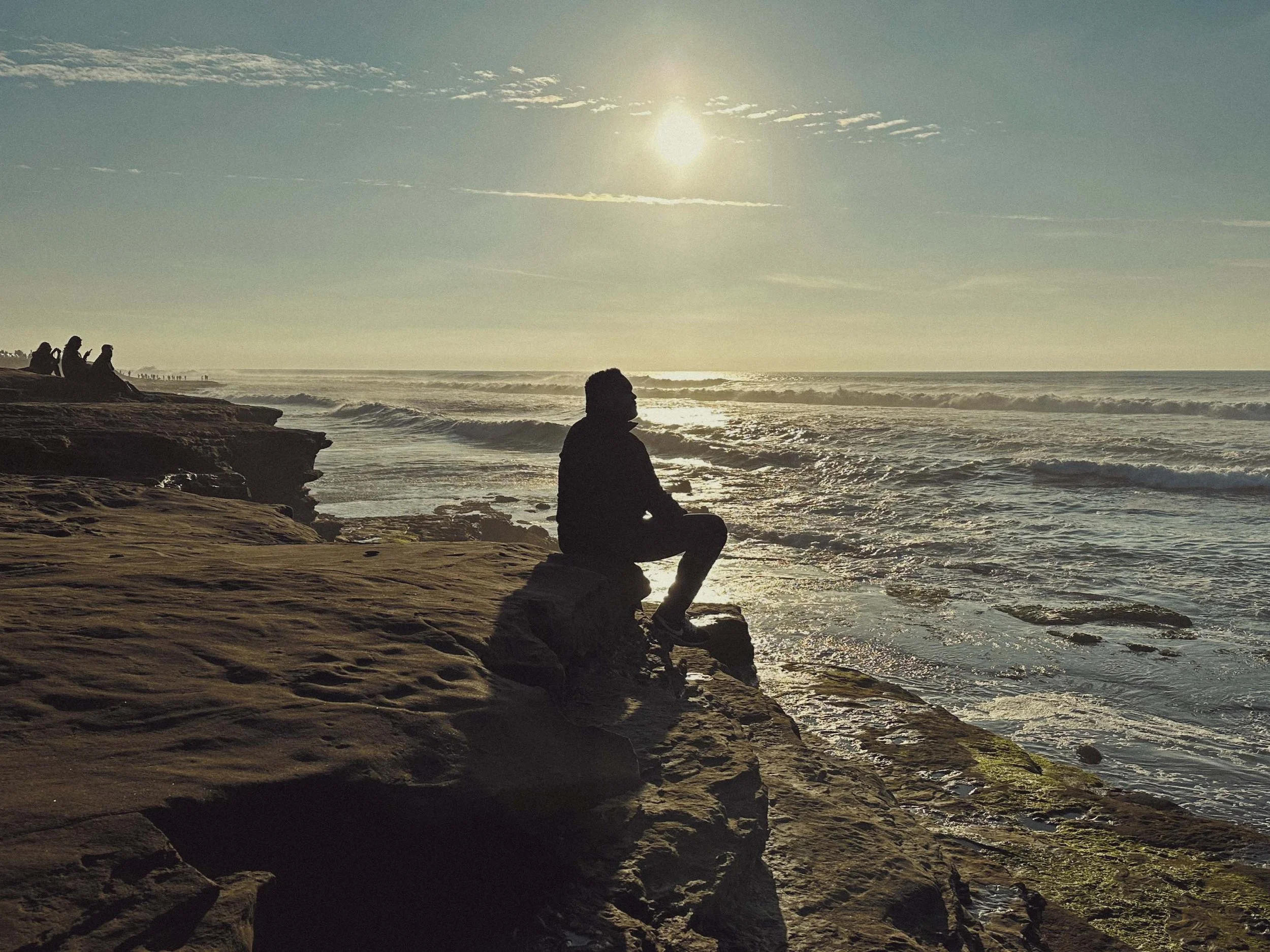 People sitting on rocks by the ocean at sunset or sunrise, with one person in the foreground in a meditation or reflective pose.