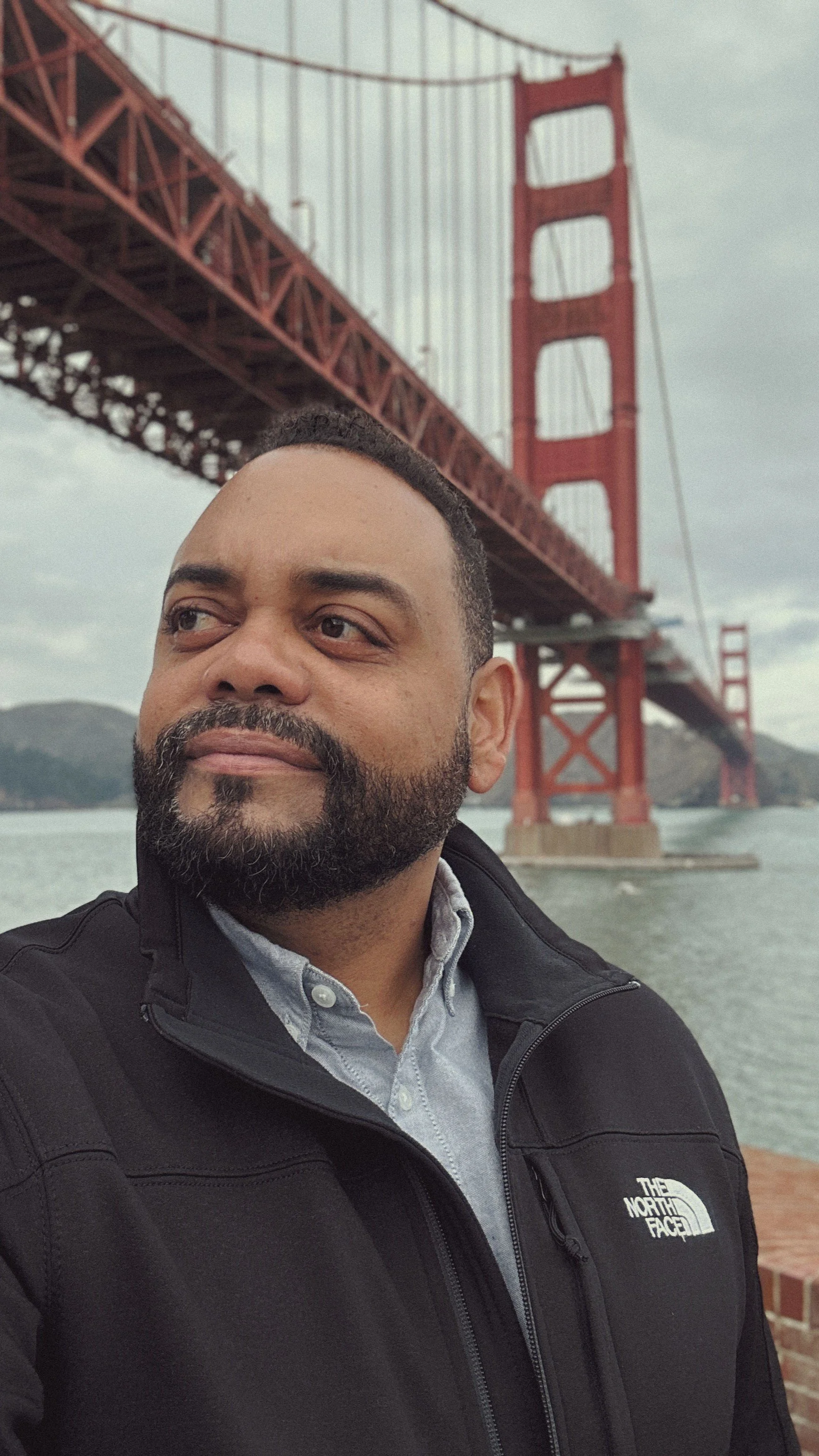 A man taking a selfie in front of the Golden Gate Bridge in San Francisco, California, with water and hills in the background.