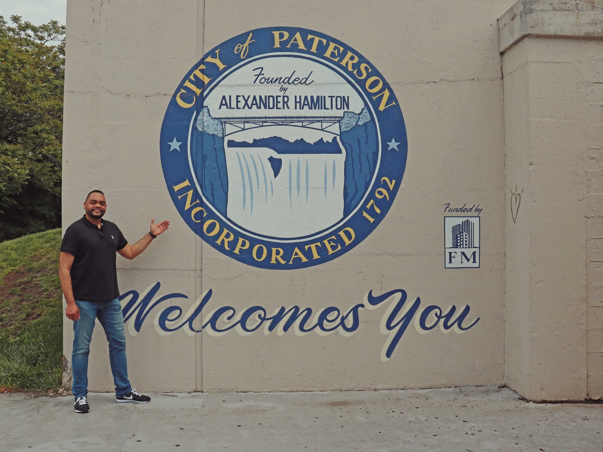 elcome sign with the city of Paterson logo and text. The sign features a graphic of a bridge and waterfall, and reads, 'City of Paterson, Founded by Alexander Hamilton, Incorporated 1792, Welcomes You.' The man is smiling and dressed in a black shirt