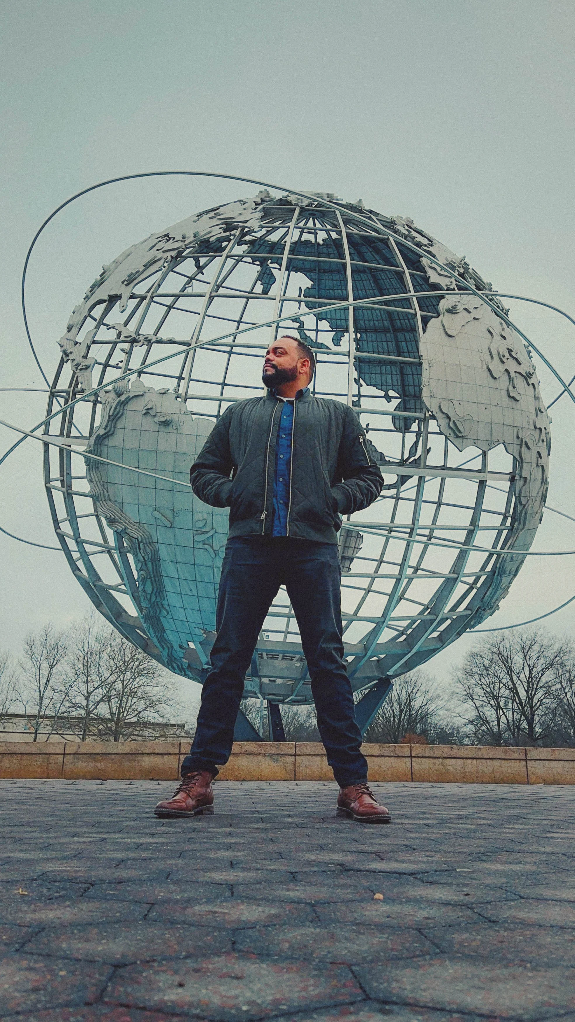A man standing with hands in pockets in front of a large globe sculpture outdoors.