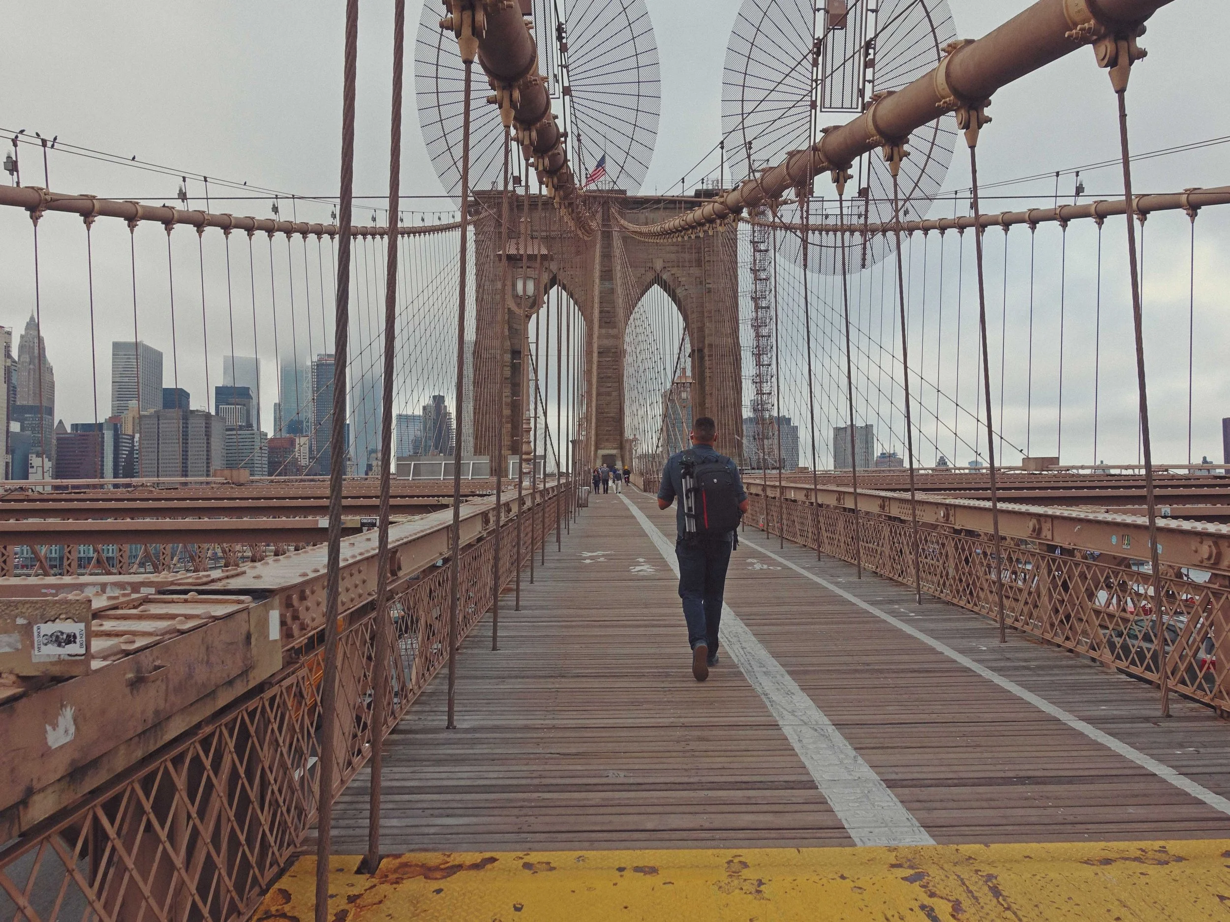 View of the Brooklyn Bridge with people walking and a person with a backpack, city skyline in the background, grey skies, and cable structures of the bridge.