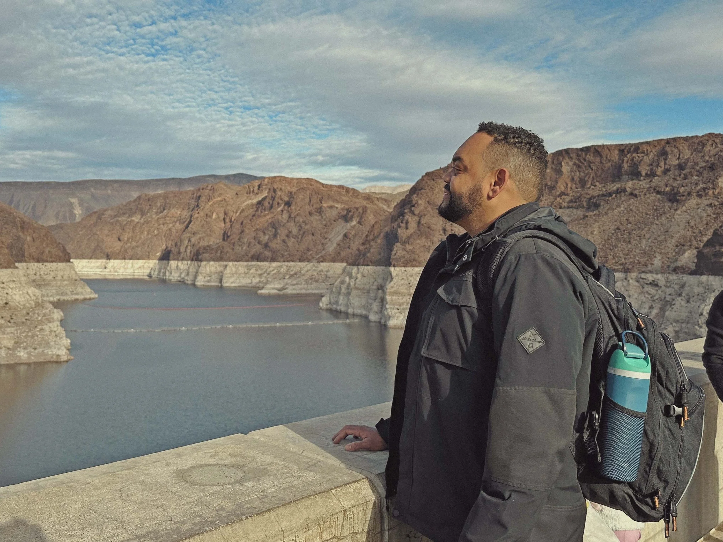 A man wearing a black jacket and a backpack stands near a concrete railing, looking to the right. Behind him is a body of water with a dam, and rugged mountains under a blue sky with scattered clouds.