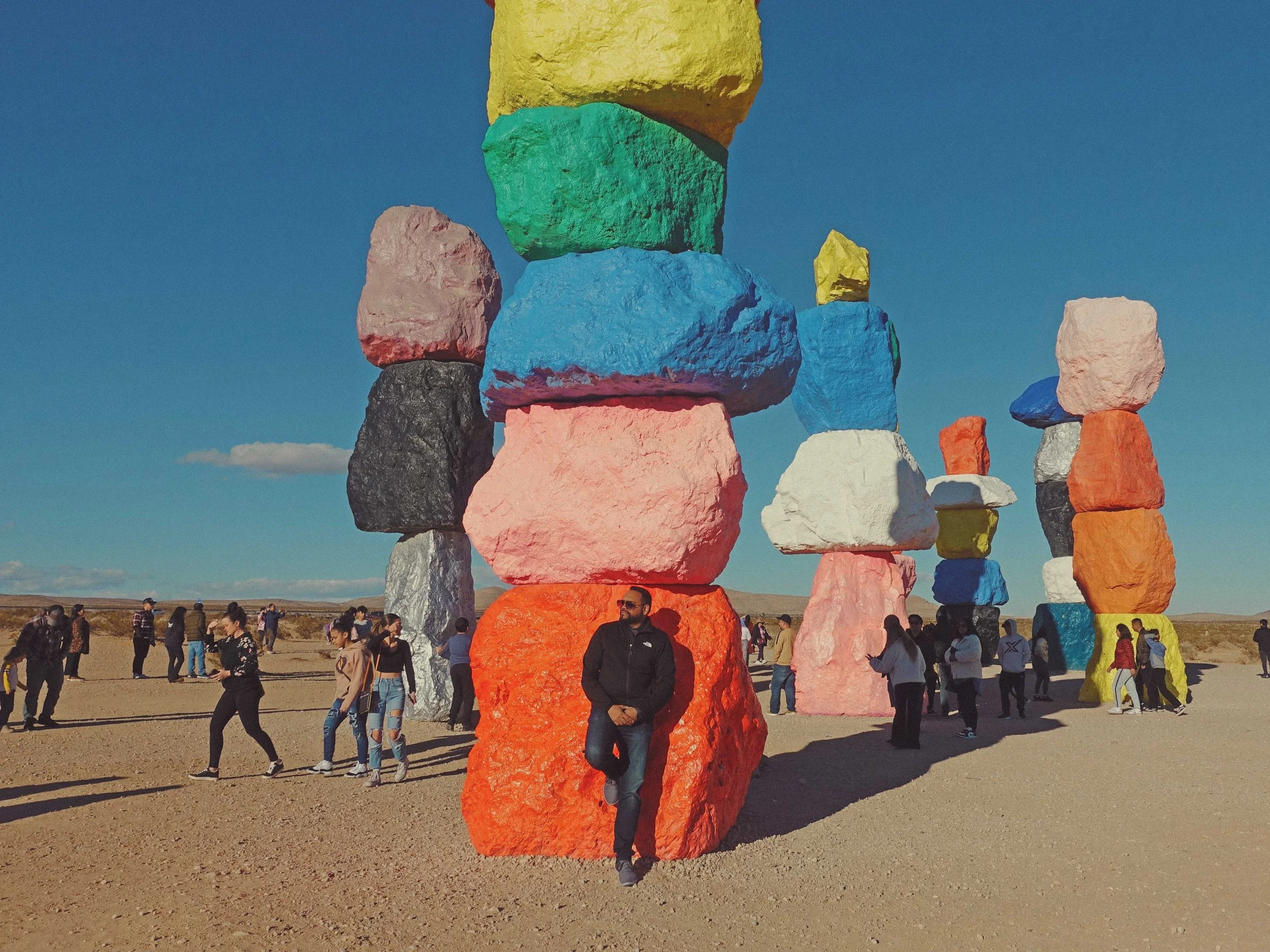 Colorful stacked rocks on a sandy landscape with people walking around, against a blue sky.