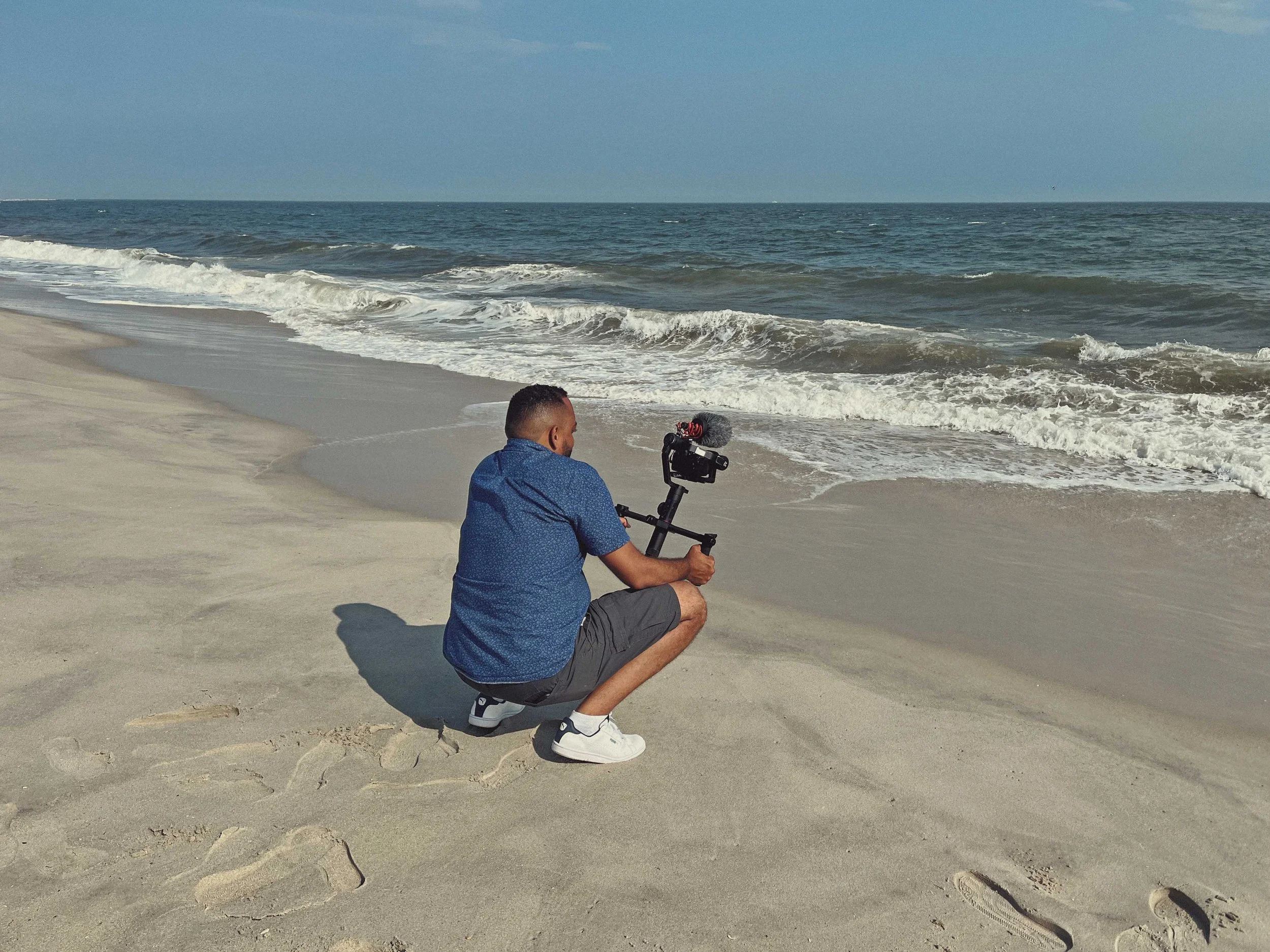 A man squatting on a sandy beach with ocean waves in the background, holding a camera on a tripod.