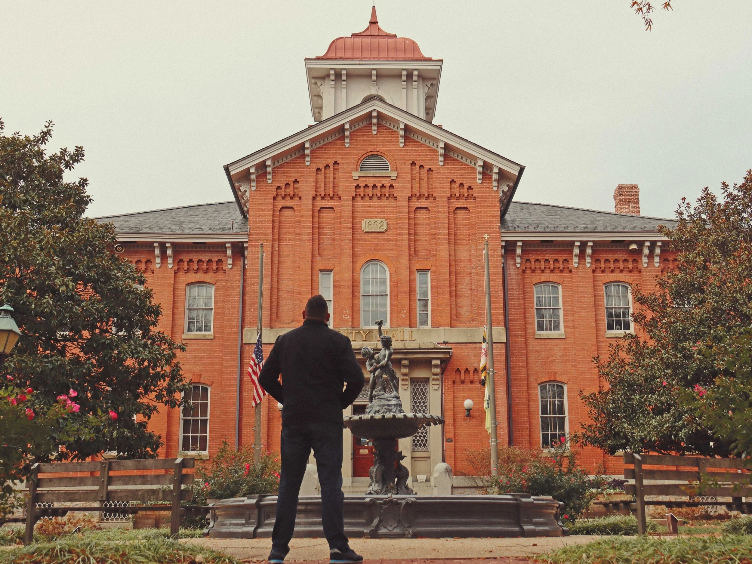 A man stands in front of a historic red brick city hall building, with a fountain in the foreground and trees on either side.