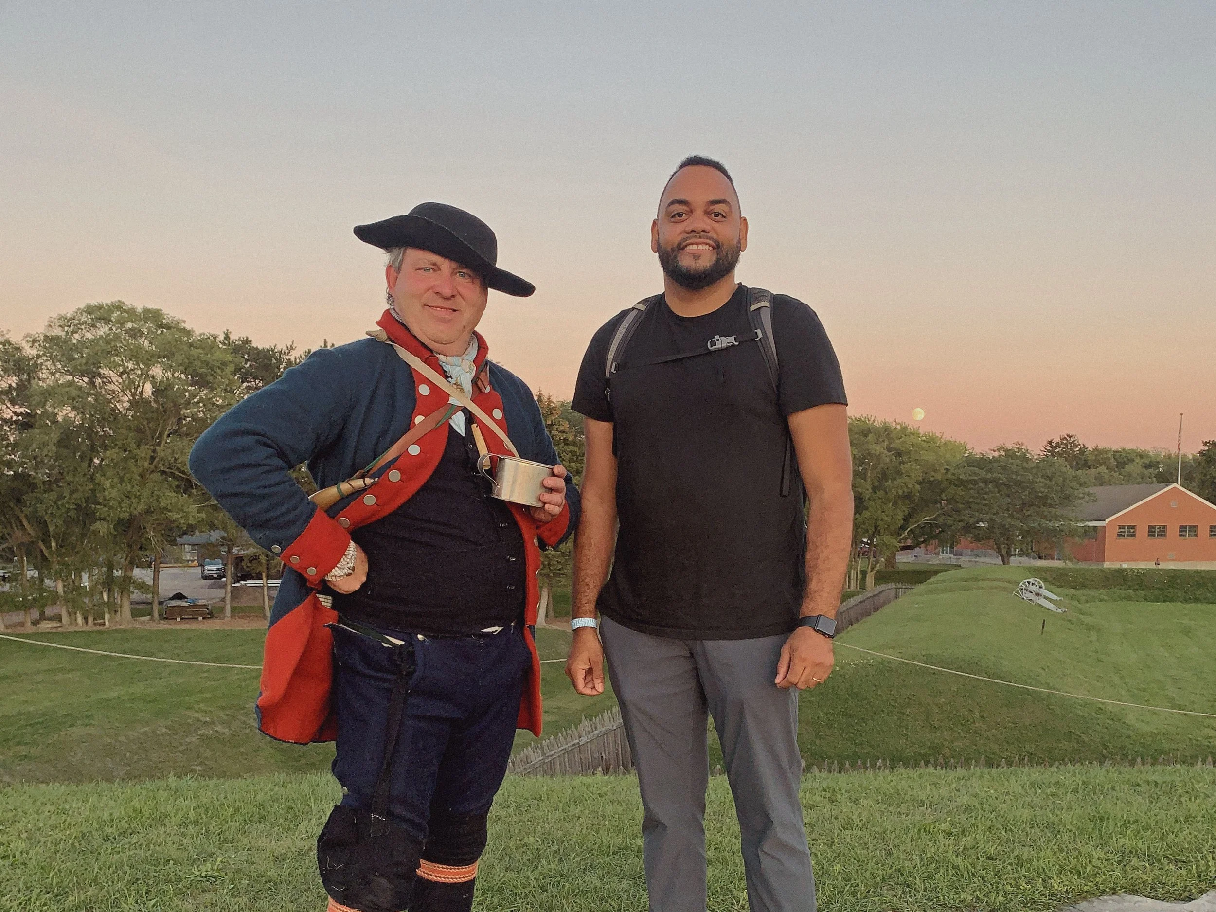 Two men standing outside on grass at sunset. One man is wearing a black hat and red jacket, holding a cup, the other is in a black shirt with a backpack. They are smiling, with trees and houses in the background.