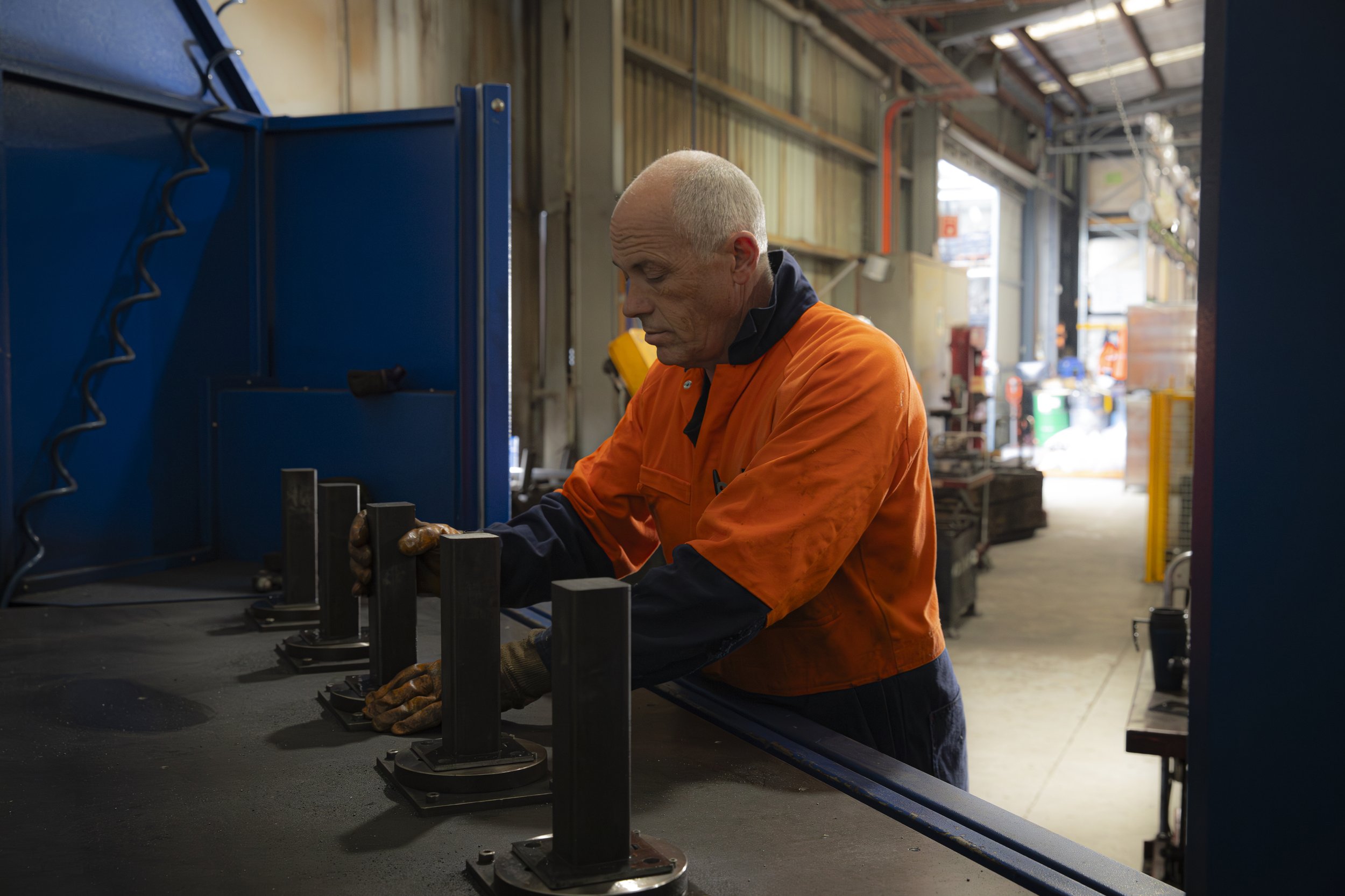 A worker in an orange safety jacket and gloves working in an industrial warehouse, handling metal parts at a workstation.