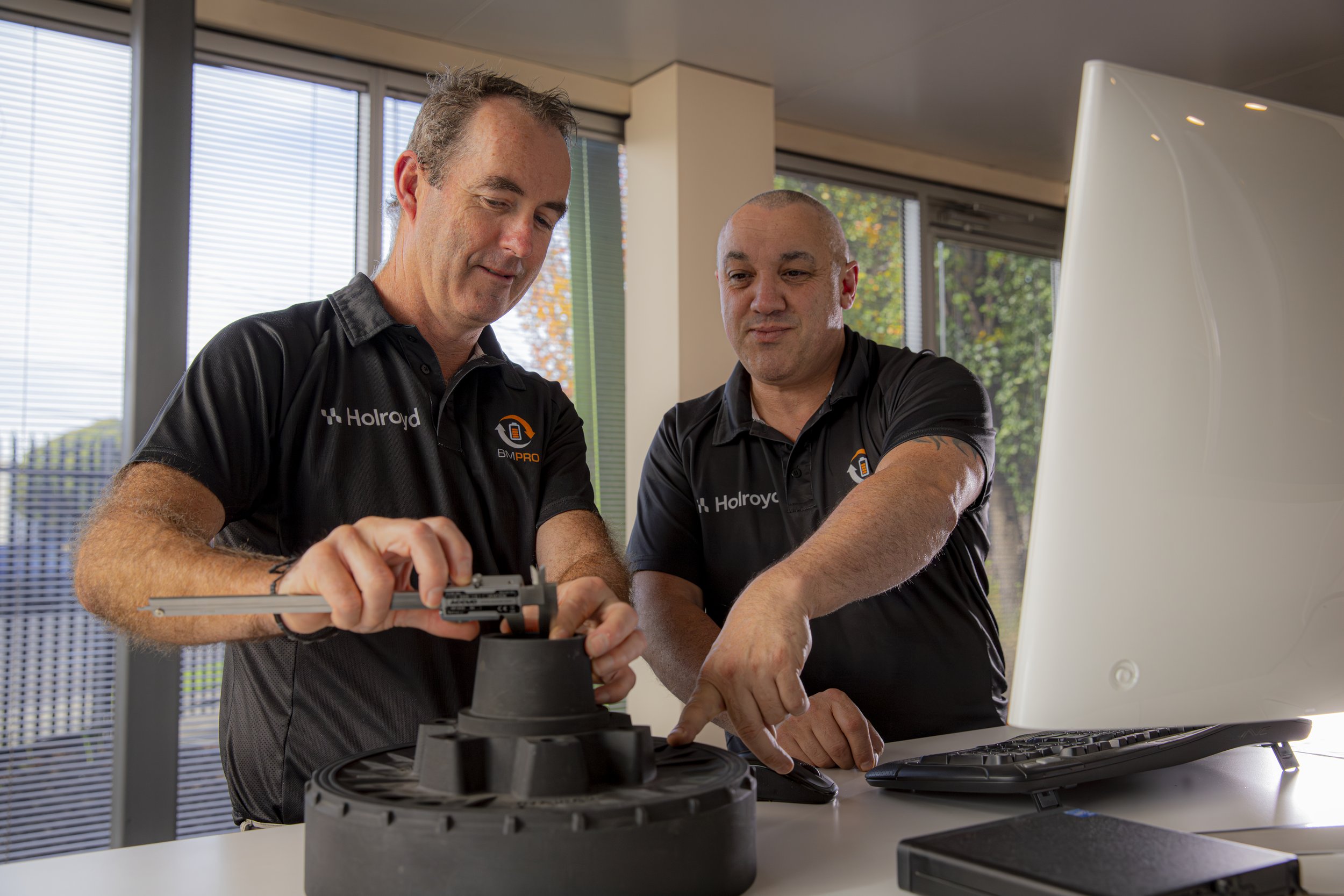 Two men wearing black polo shirts working together on a tire, with one using a wrench, in a modern office with large windows.