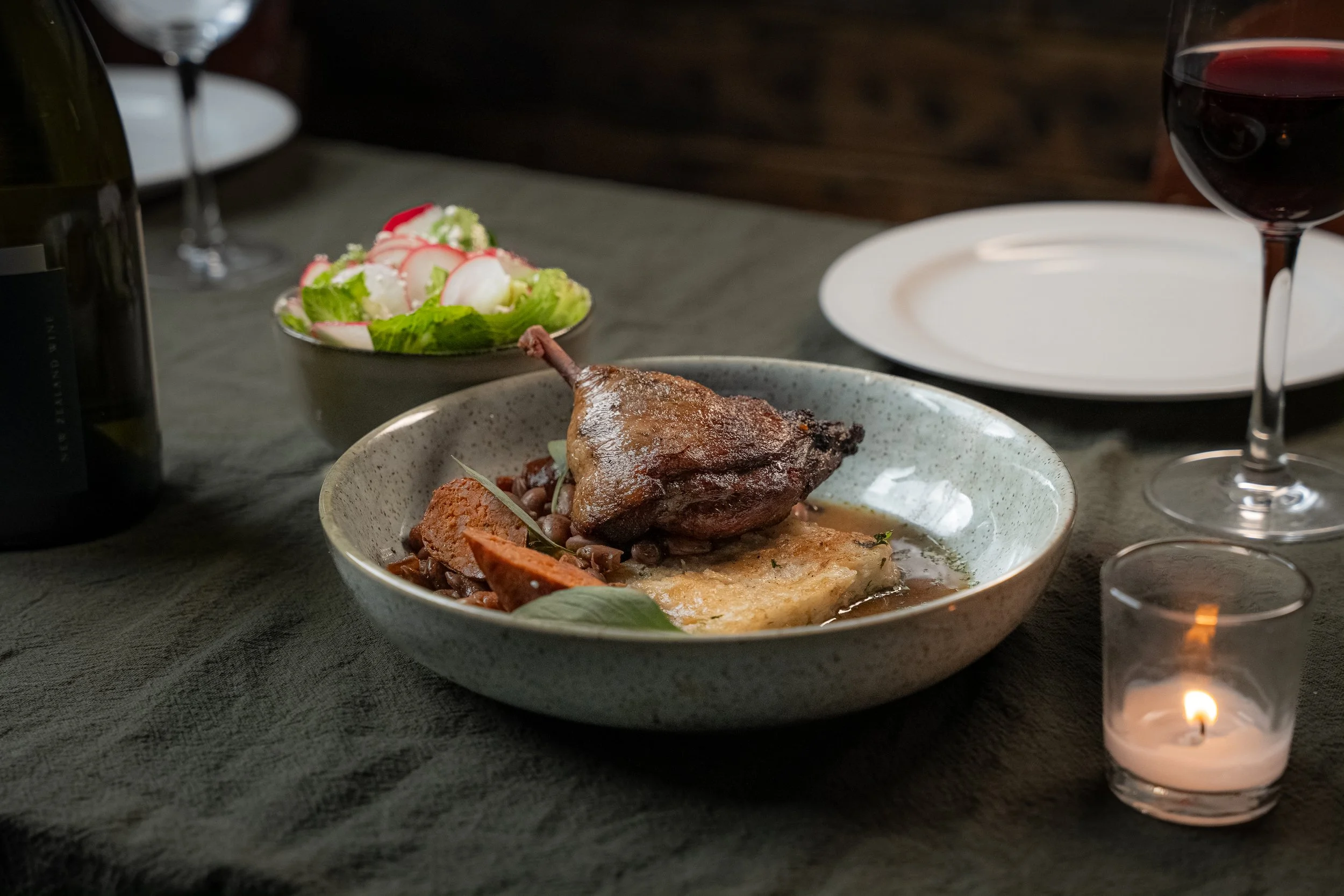 Plate of cooked duck with sauce, vegetables, and lentils on a dining table, with a side salad in a bowl, wine glass of red wine, unfilled white plate, and a lit candle.