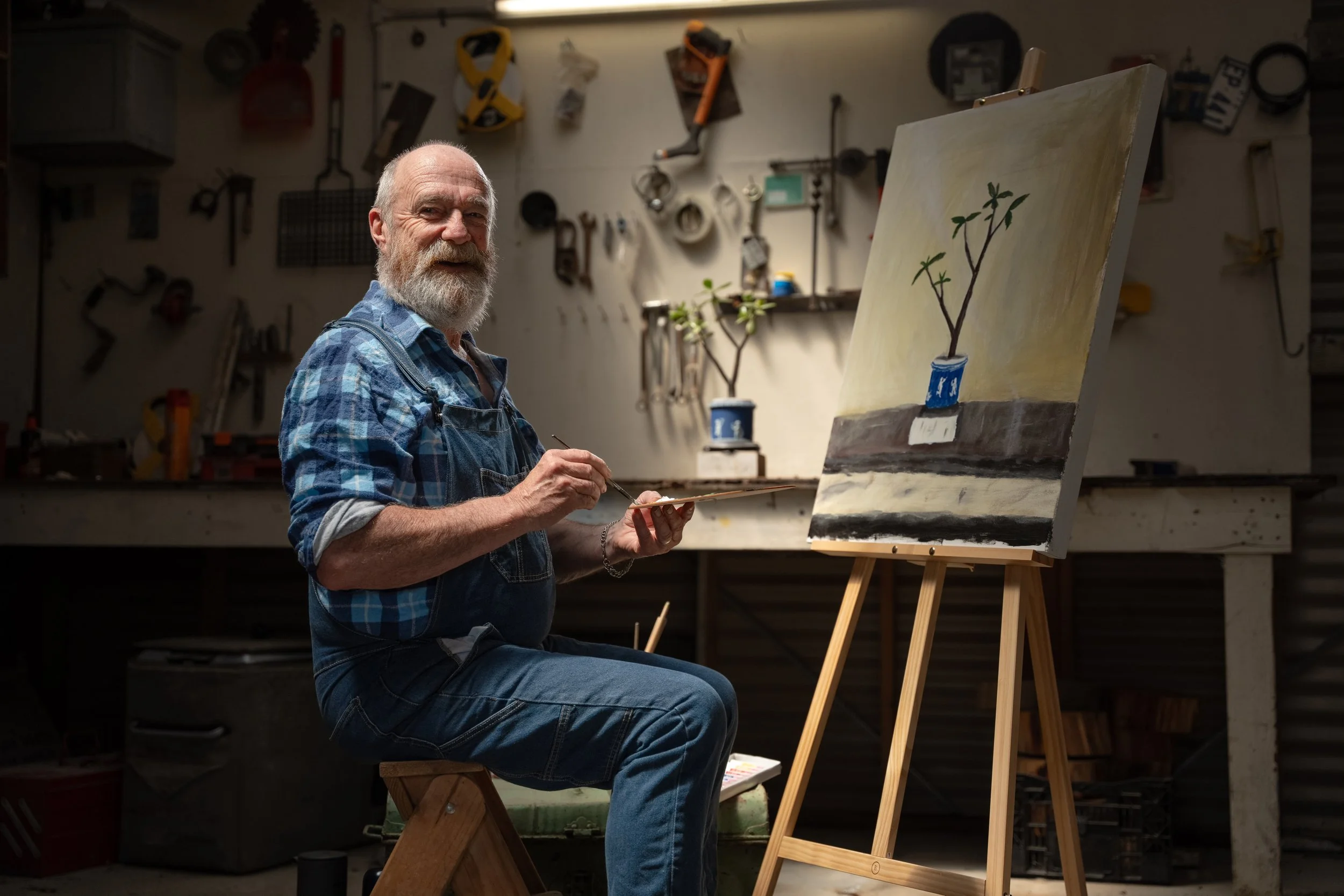 An elderly man with a beard, wearing a plaid shirt and denim overalls, sitting on a stool in a workshop, painting a picture of a potted plant on a canvas. The workshop background features various tools hanging on the wall.