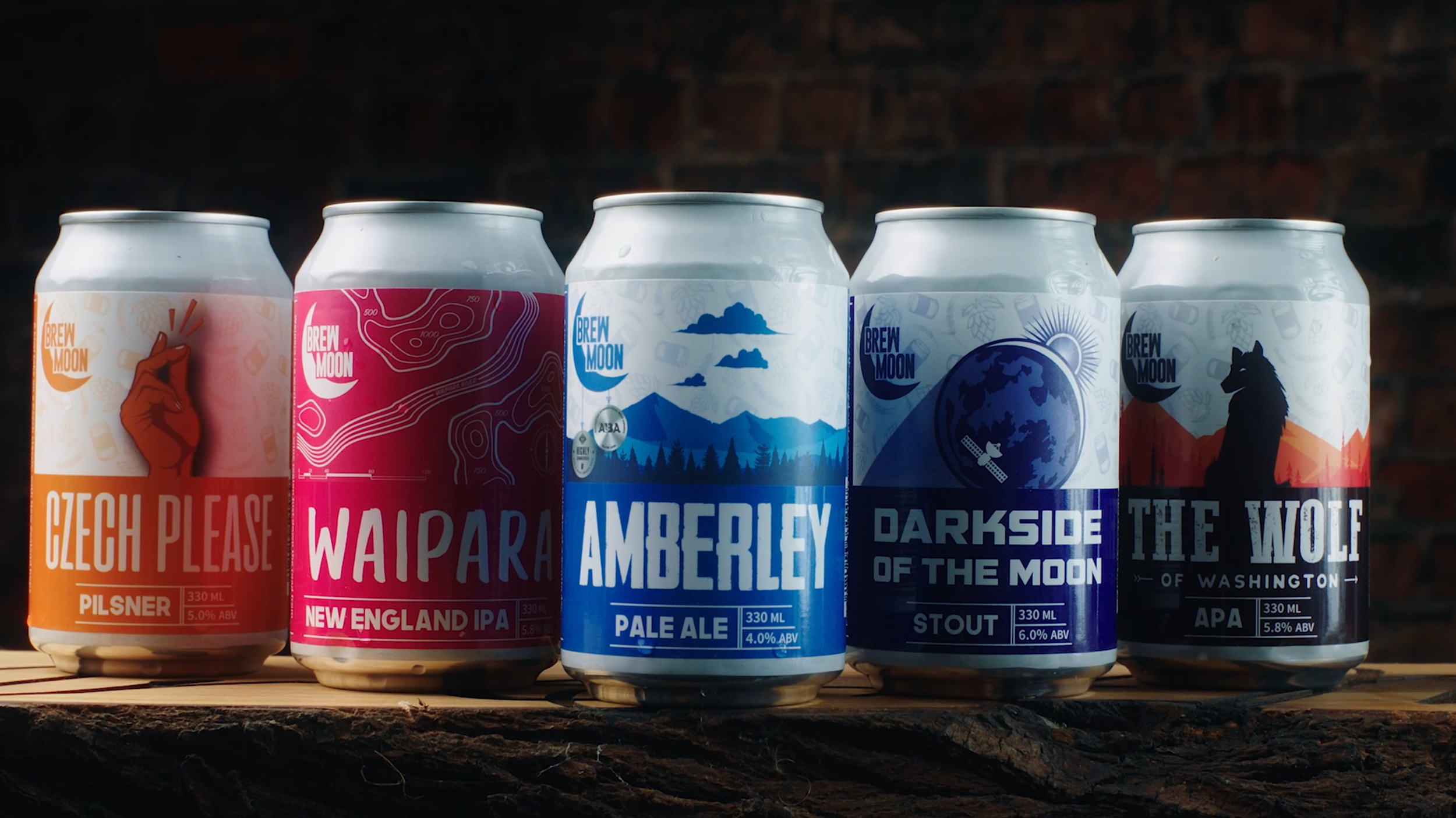 Five cans of Brew Moon beer, each with unique labels, arranged in a row on a wooden surface against a brick wall background.
