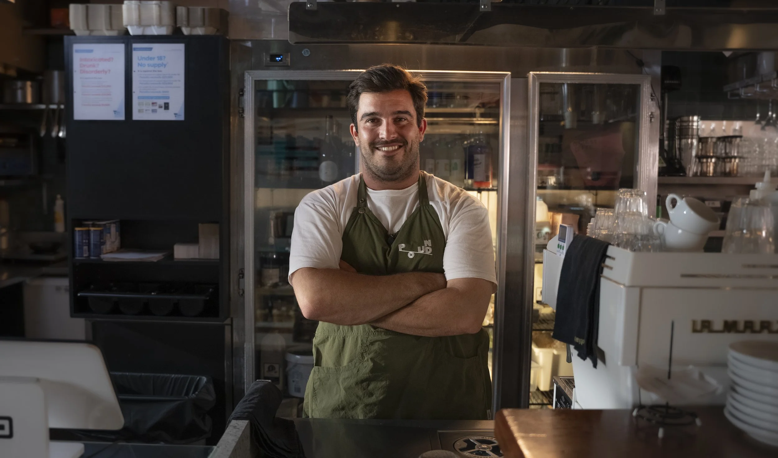 A smiling man with dark hair and a beard, wearing a white t-shirt and an olive green apron, stands with arms crossed in a kitchen or cafe setting, behind a glass door refrigerator and some kitchen appliances.