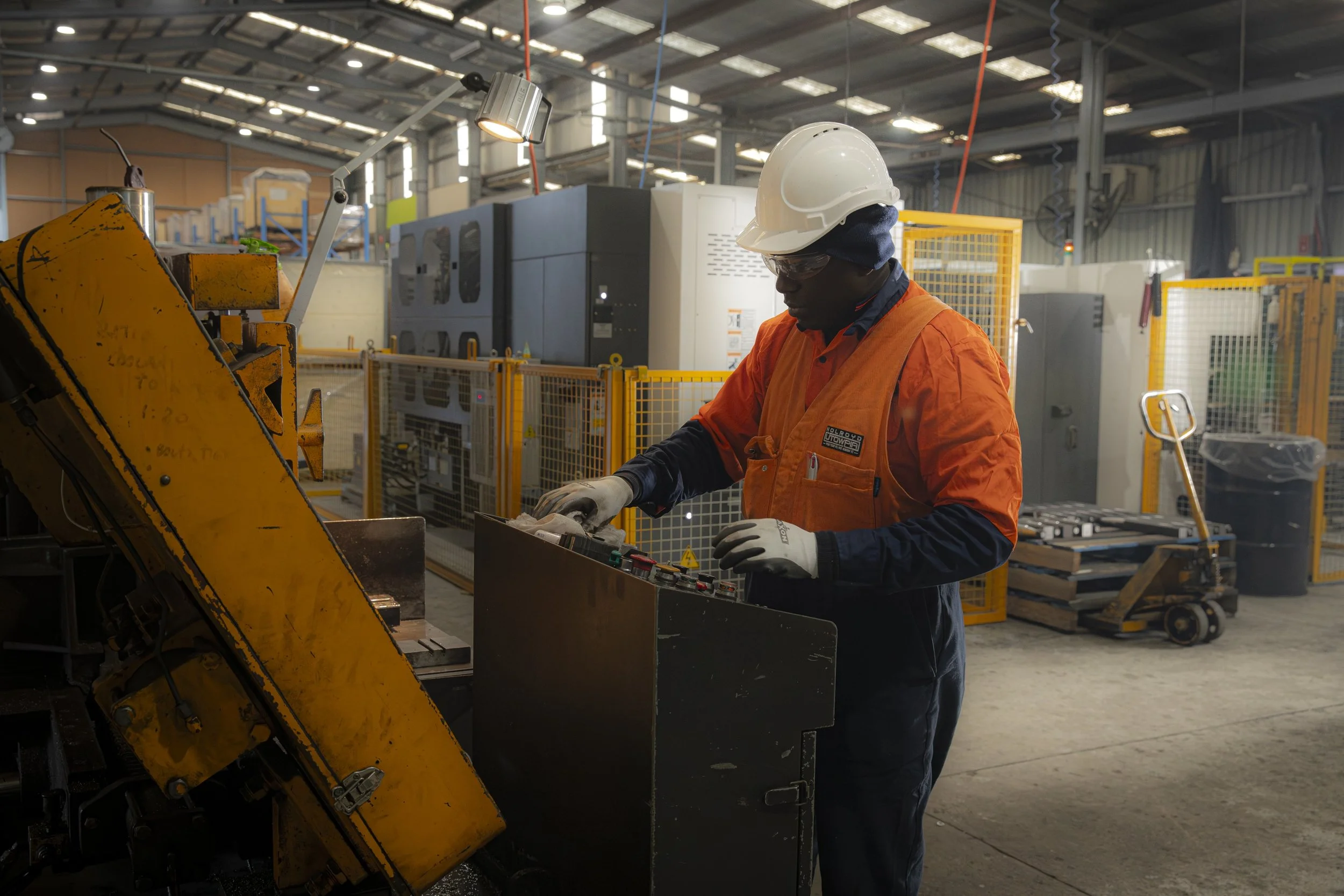 An industrial worker wearing safety gear operating a machine inside a factory or workshop with various machinery and equipment in the background.