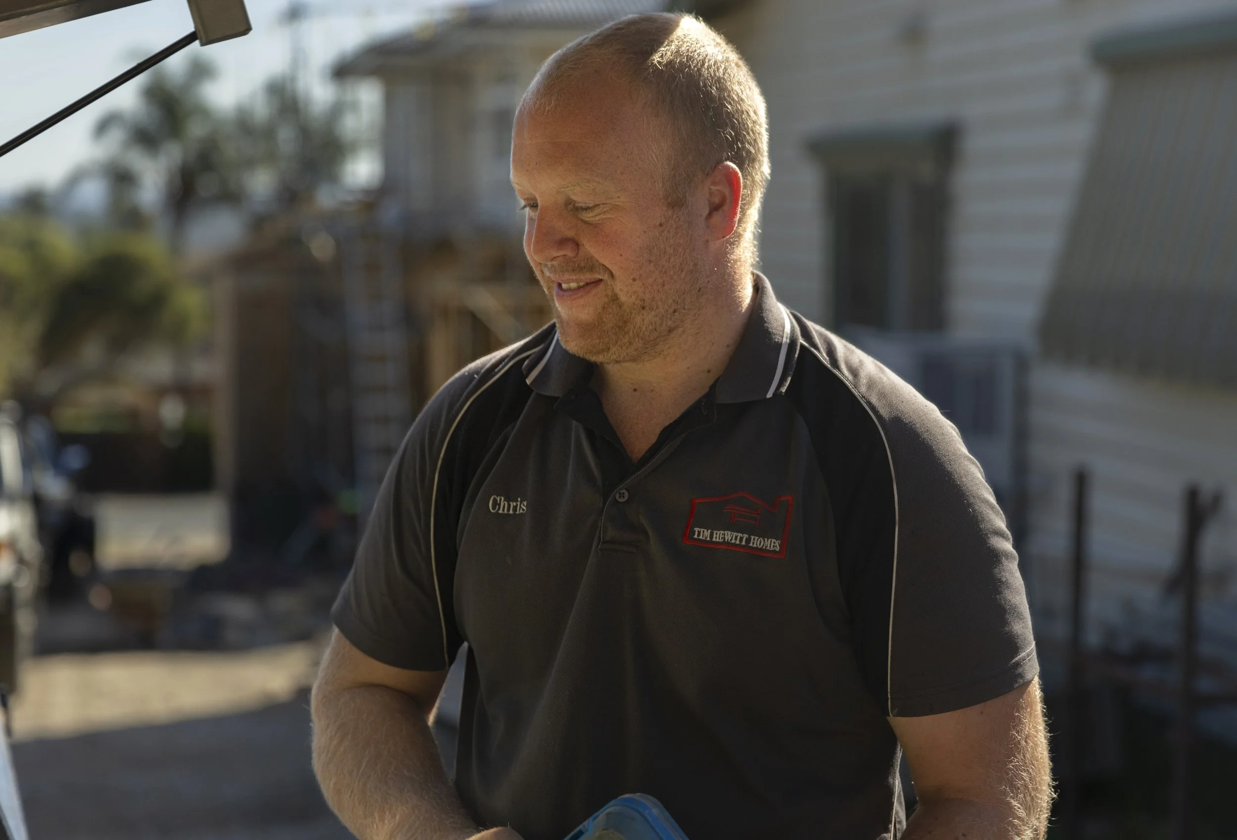 A man with short blond hair wearing a black polo shirt with logos, standing outdoors beside a white house, smiling and looking down.