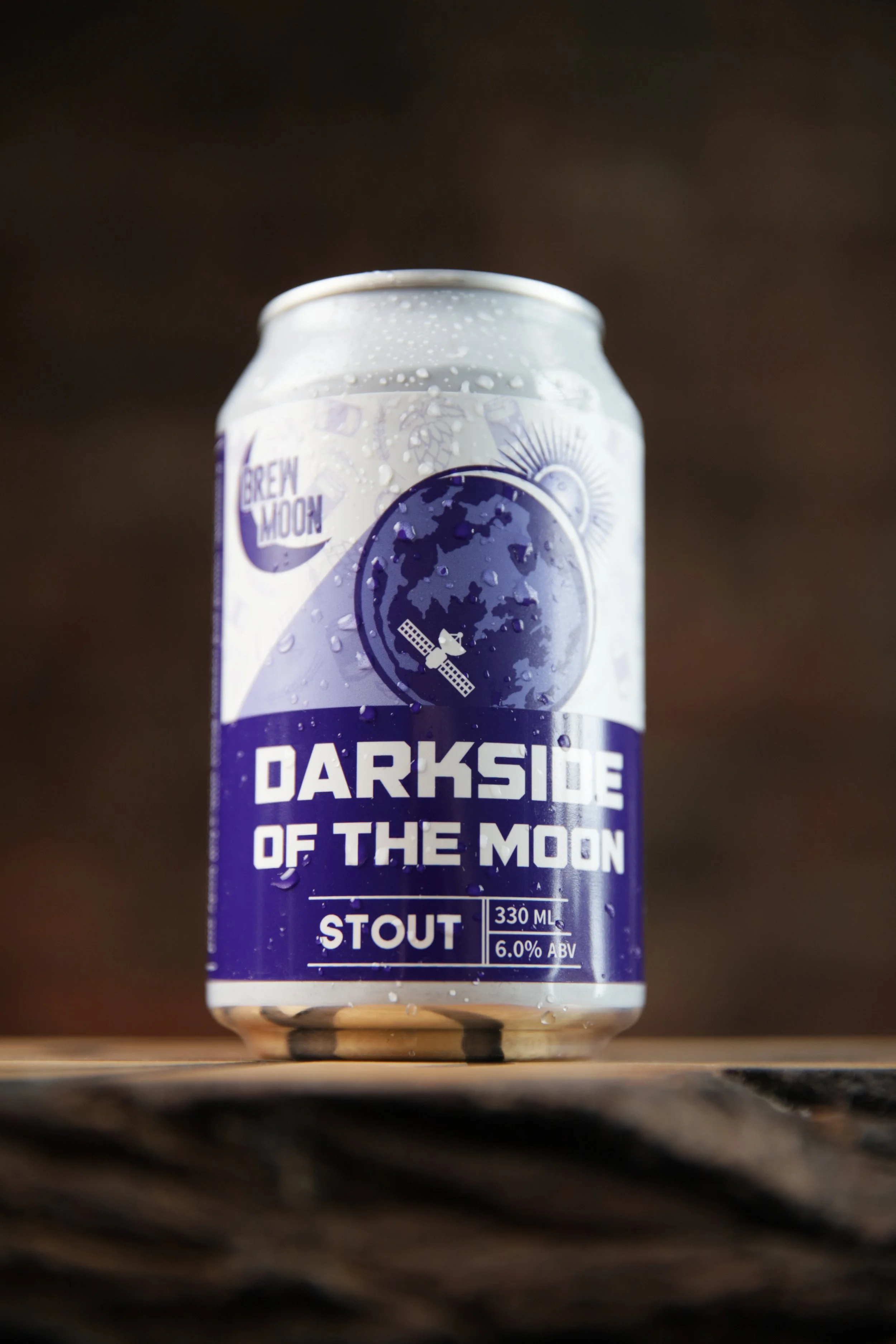 A can of Brew Moon Darkside of the Moon stout beer with a purple and white label, featuring an illustration of the moon with a satellite, placed on a wooden surface.