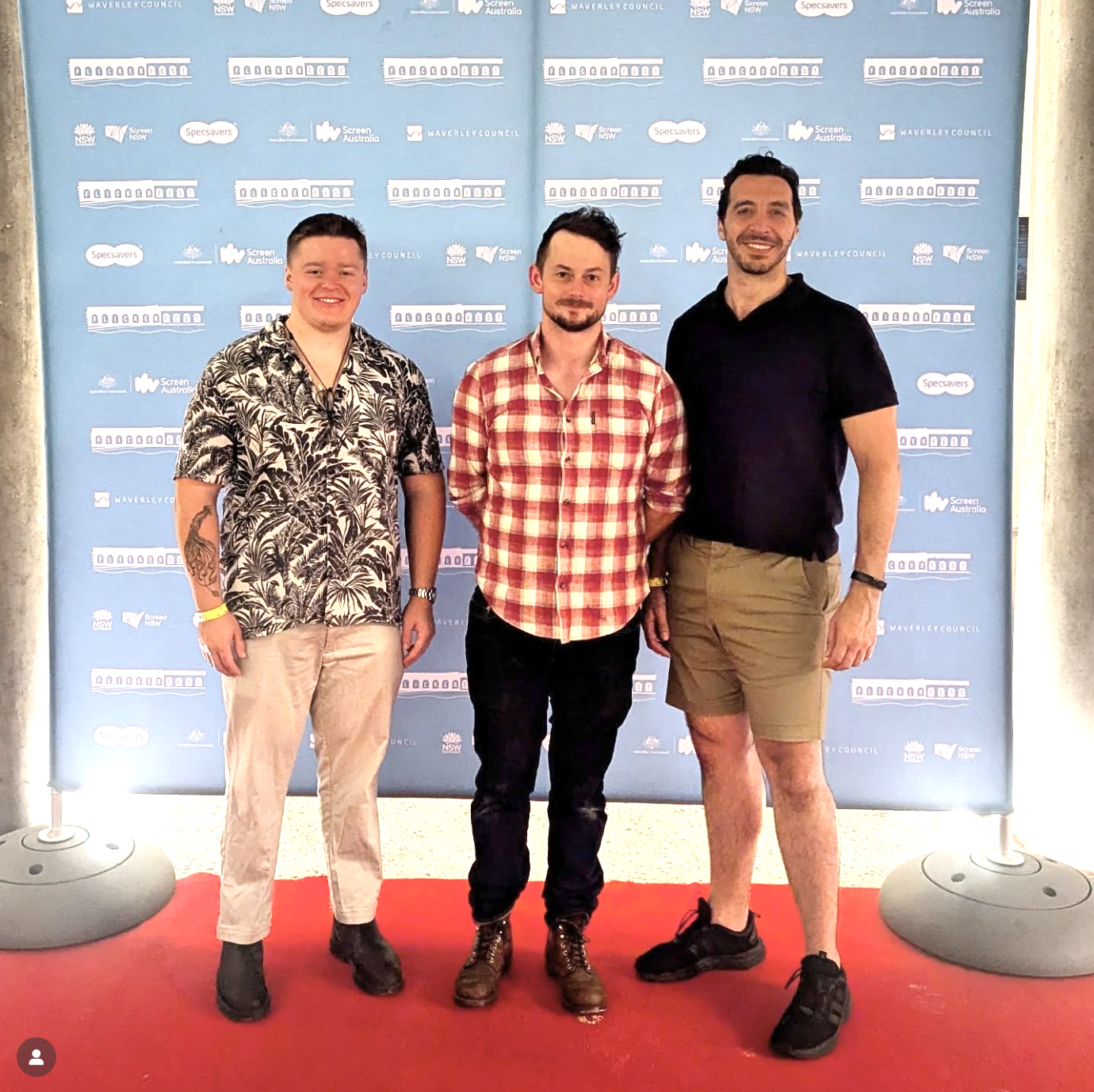 Three men standing on a red carpet in front of a blue backdrop with logos, smiling at the camera.