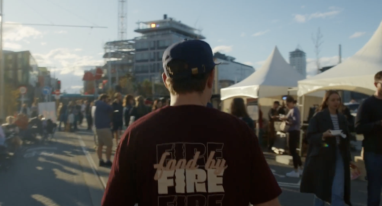 A person wearing a black cap and a maroon shirt with white text, seen from behind, walking through a busy outdoor event with tents, people, and buildings under a cloudy sky.