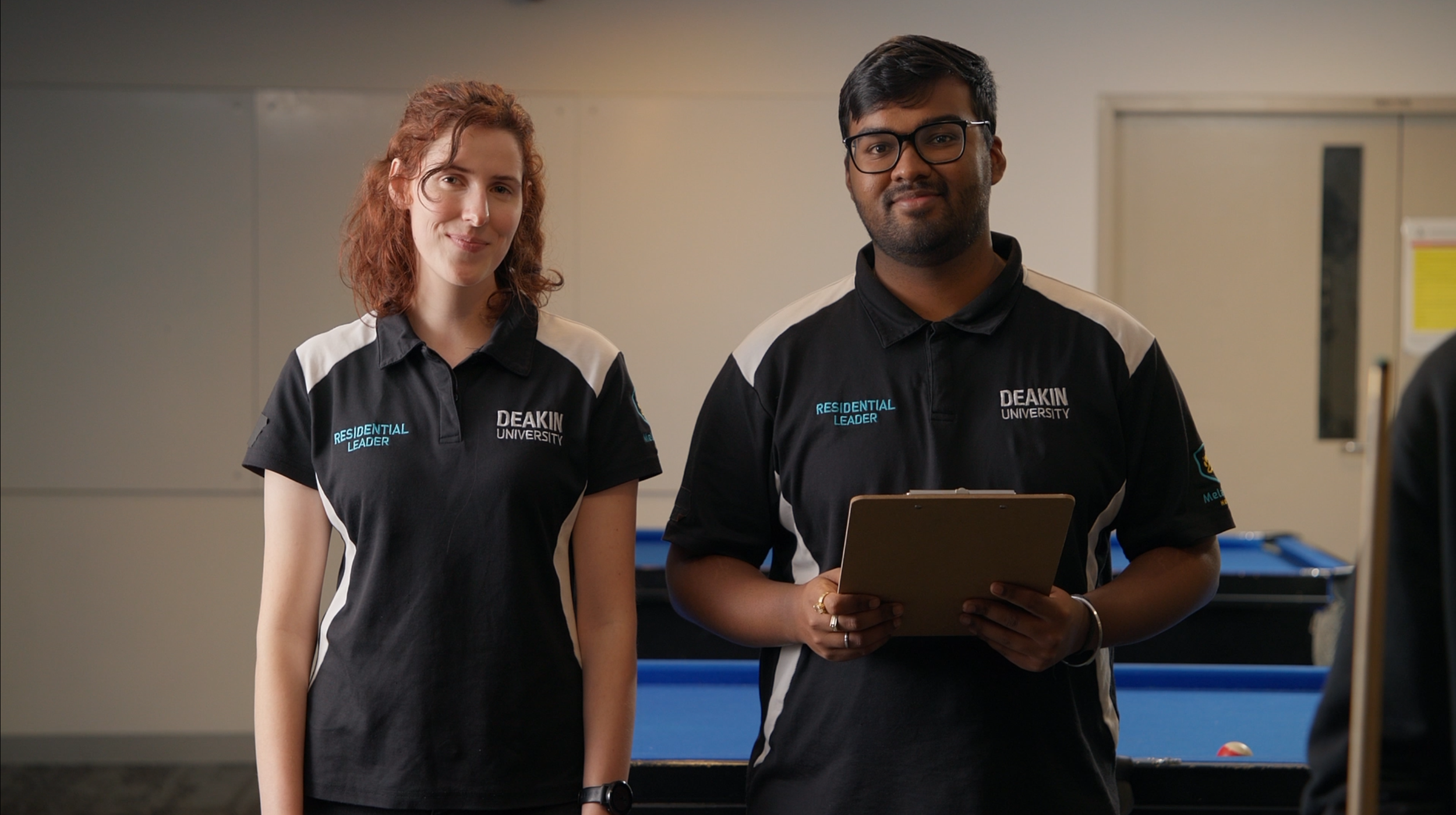 Two students wearing black Deakin University shirts with 'Resident Leader' patches, standing in a room with a pool table in the background, smiling at the camera.