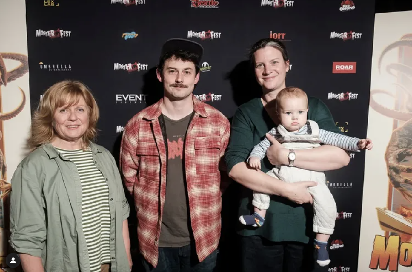 Group photo of four people, including a woman, a man with a baseball cap, a man holding a young child, standing in front of a black backdrop with logos, at Monster Fest event.