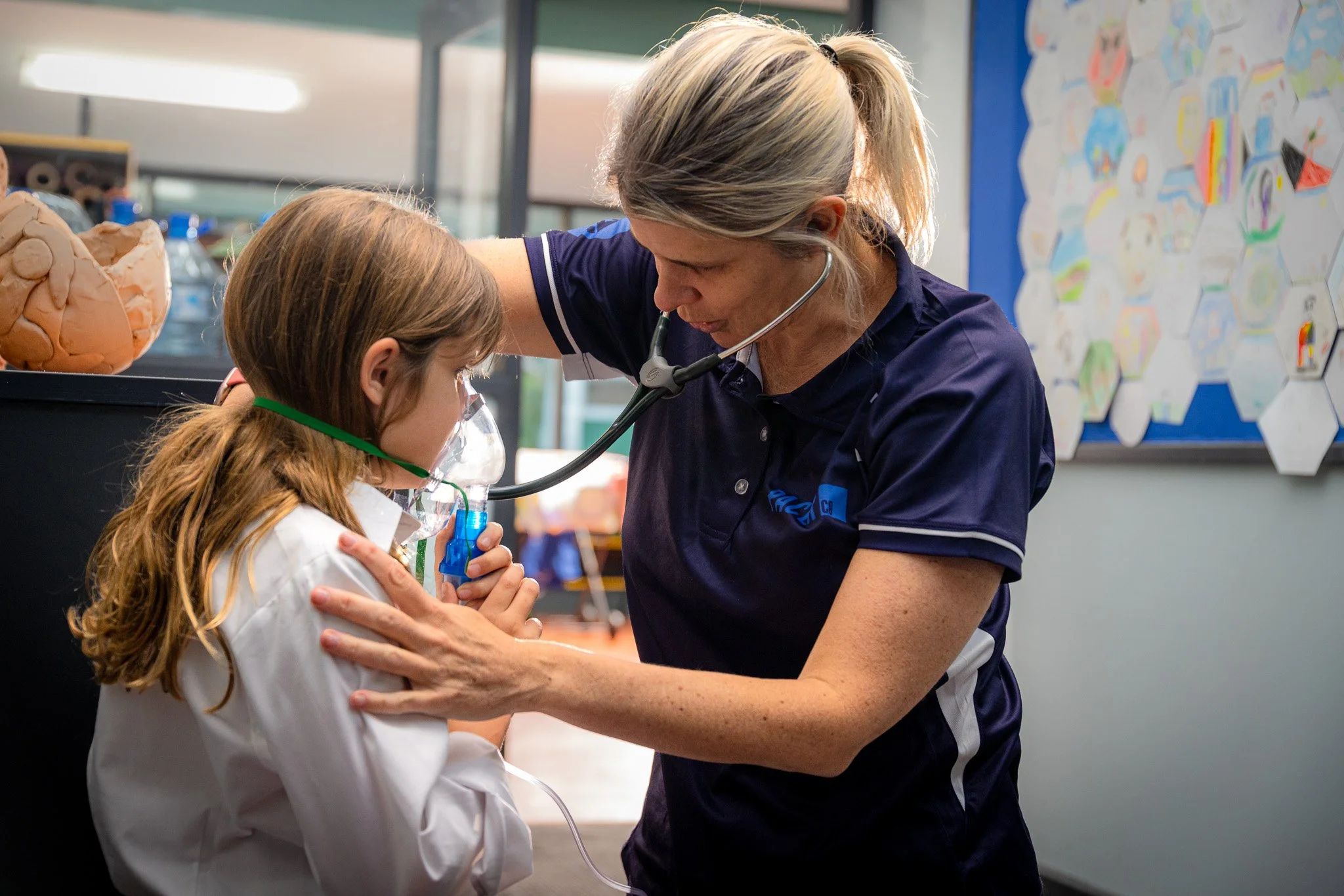 A female healthcare professional examining a young girl with a stethoscope in a classroom setting.