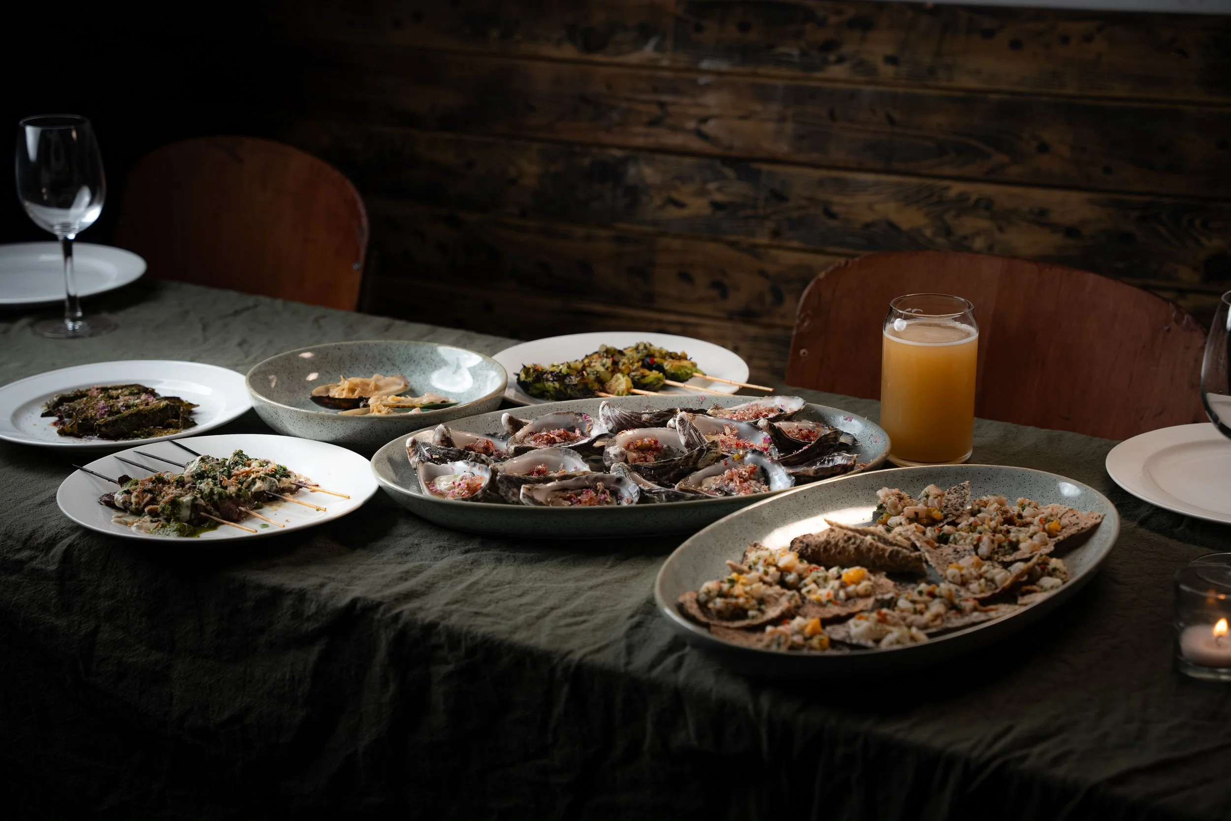 A table with various plates of food, including oysters, tacos, and grilled skewers, with a glass of orange juice and empty plates in the background.