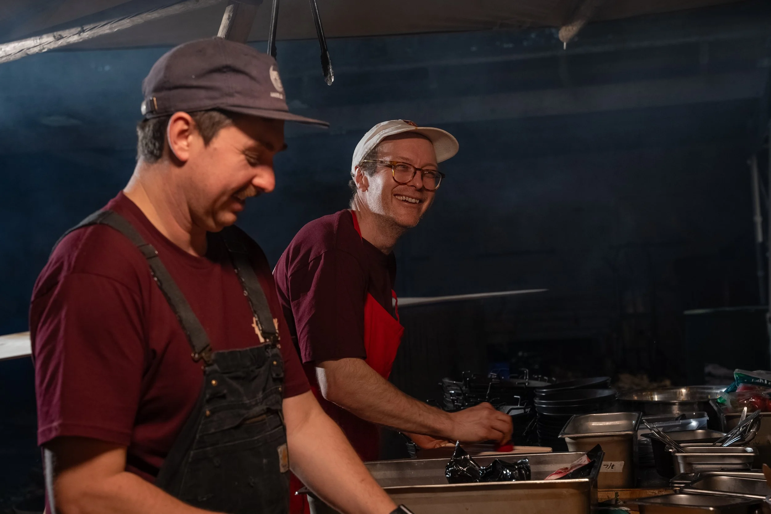 Two men preparing food in a kitchen, smiling and wearing casual shirts, one with a cap and apron.
