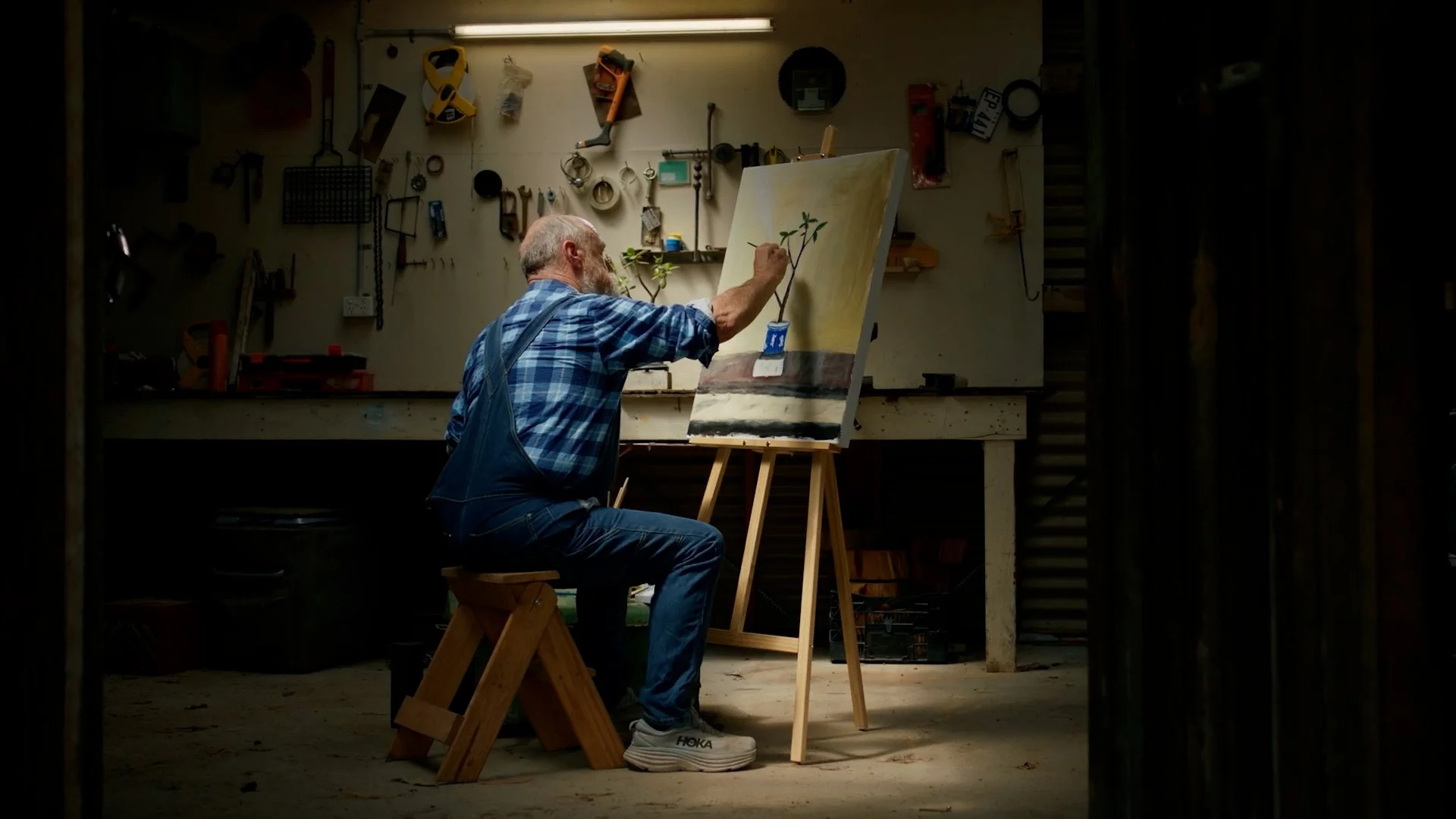 An elderly man with a gray beard painting a plant on a canvas in a workshop, seated on a small wooden stool, surrounded by various tools and equipment.