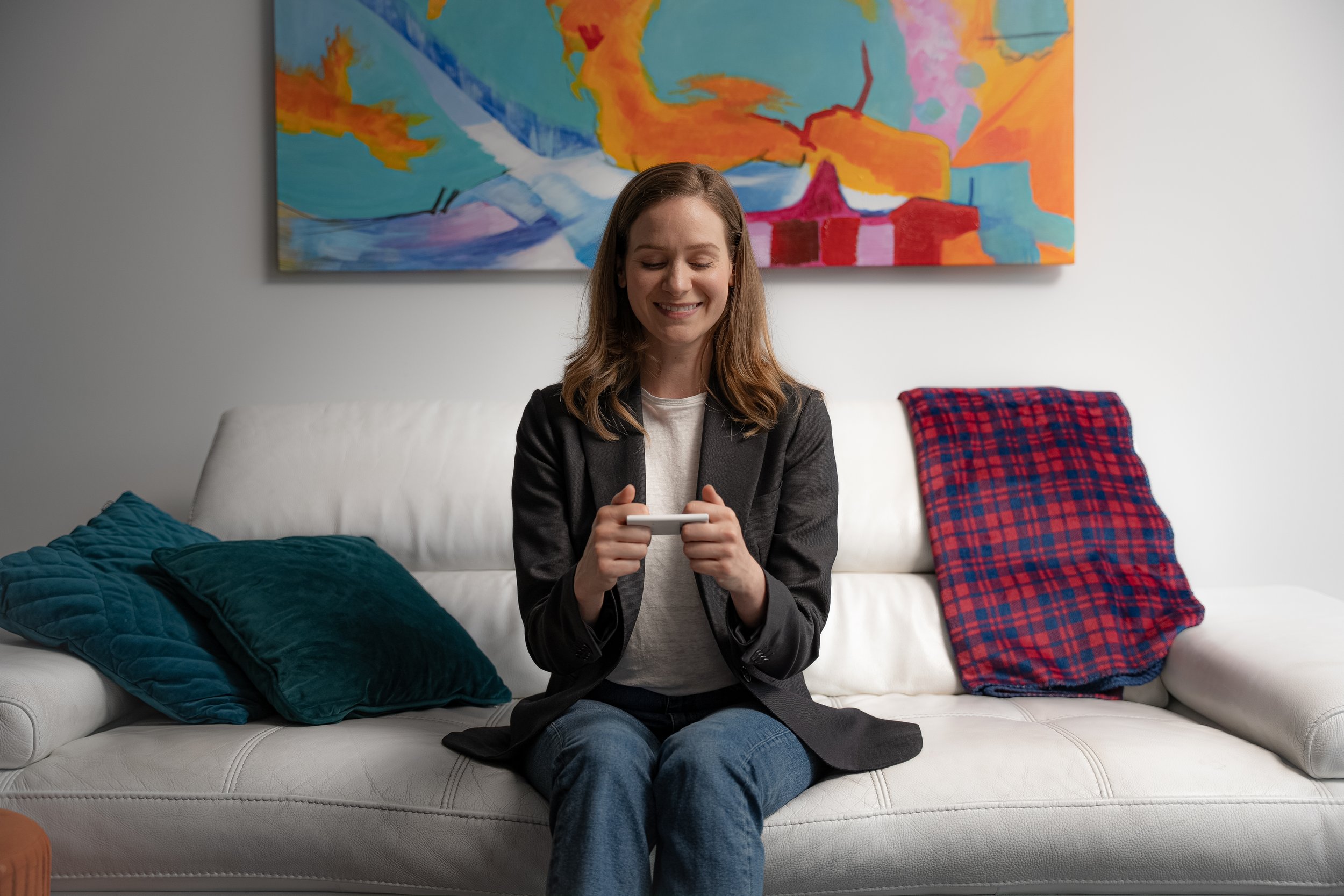 A woman with shoulder-length brown hair, wearing a black blazer and jeans, sitting on a white sofa, smiling while looking at her phone. The sofa has a teal plush pillow and a red and blue checkered blanket on it. A colorful abstract painting hangs on