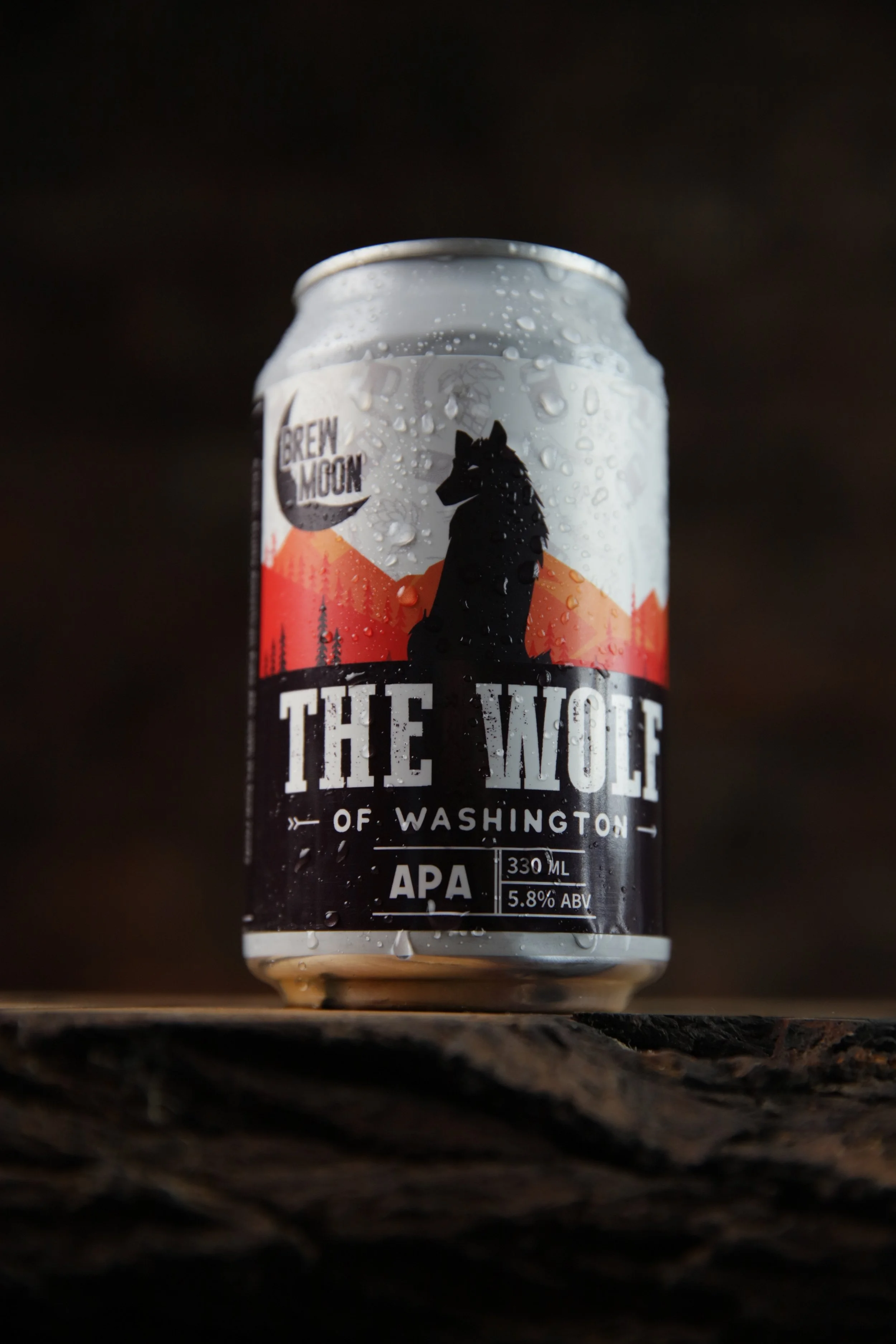 A can of Brew Moon beer called 'The Wolf of Washington,' featuring a black silhouette of a wolf against a red and orange mountain landscape with trees, water droplets on the can, and a dark blurred background.