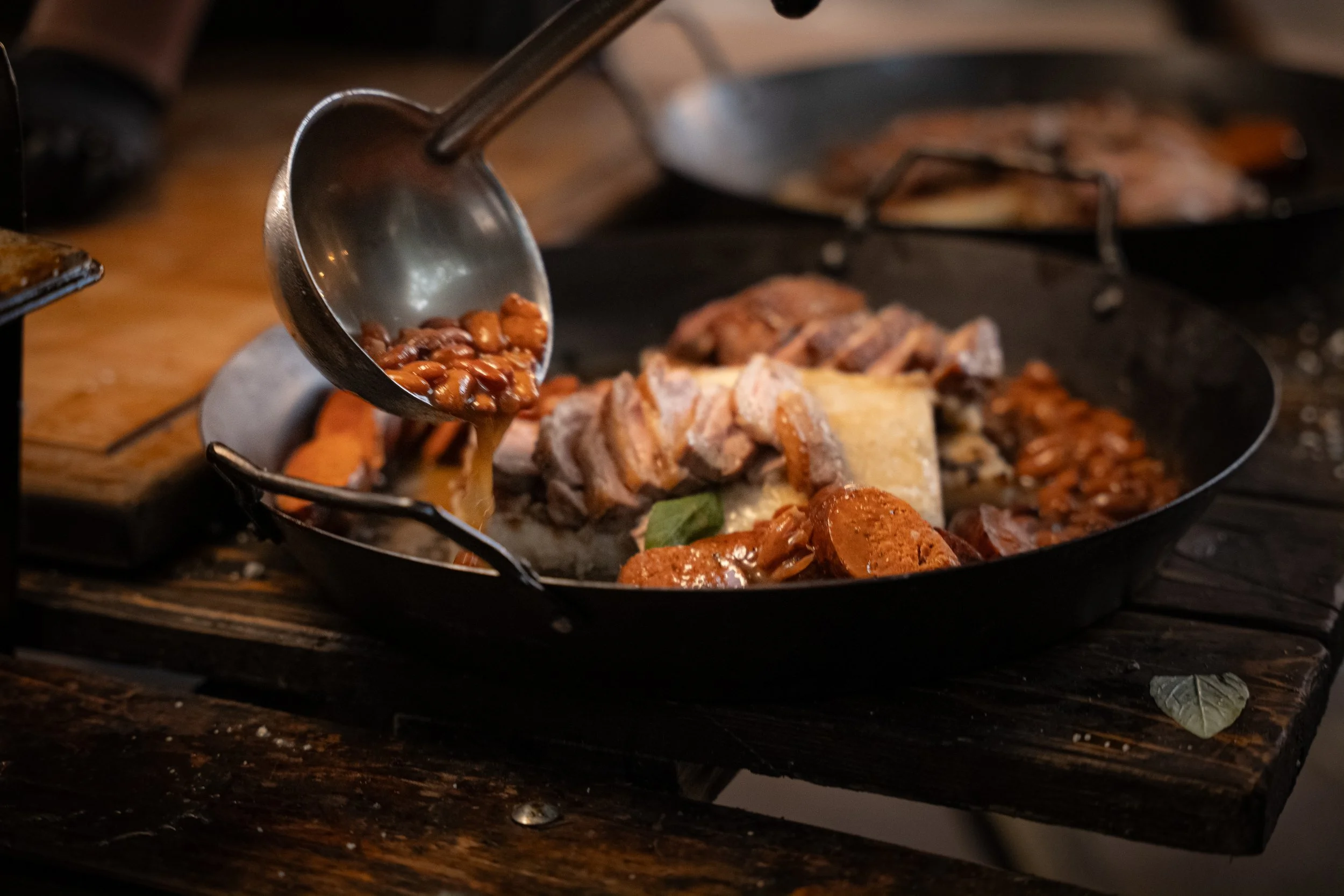 A skillet on a wooden surface with cooked meats, beans, and vegetables, as a person pours beans into the skillet from a ladle.