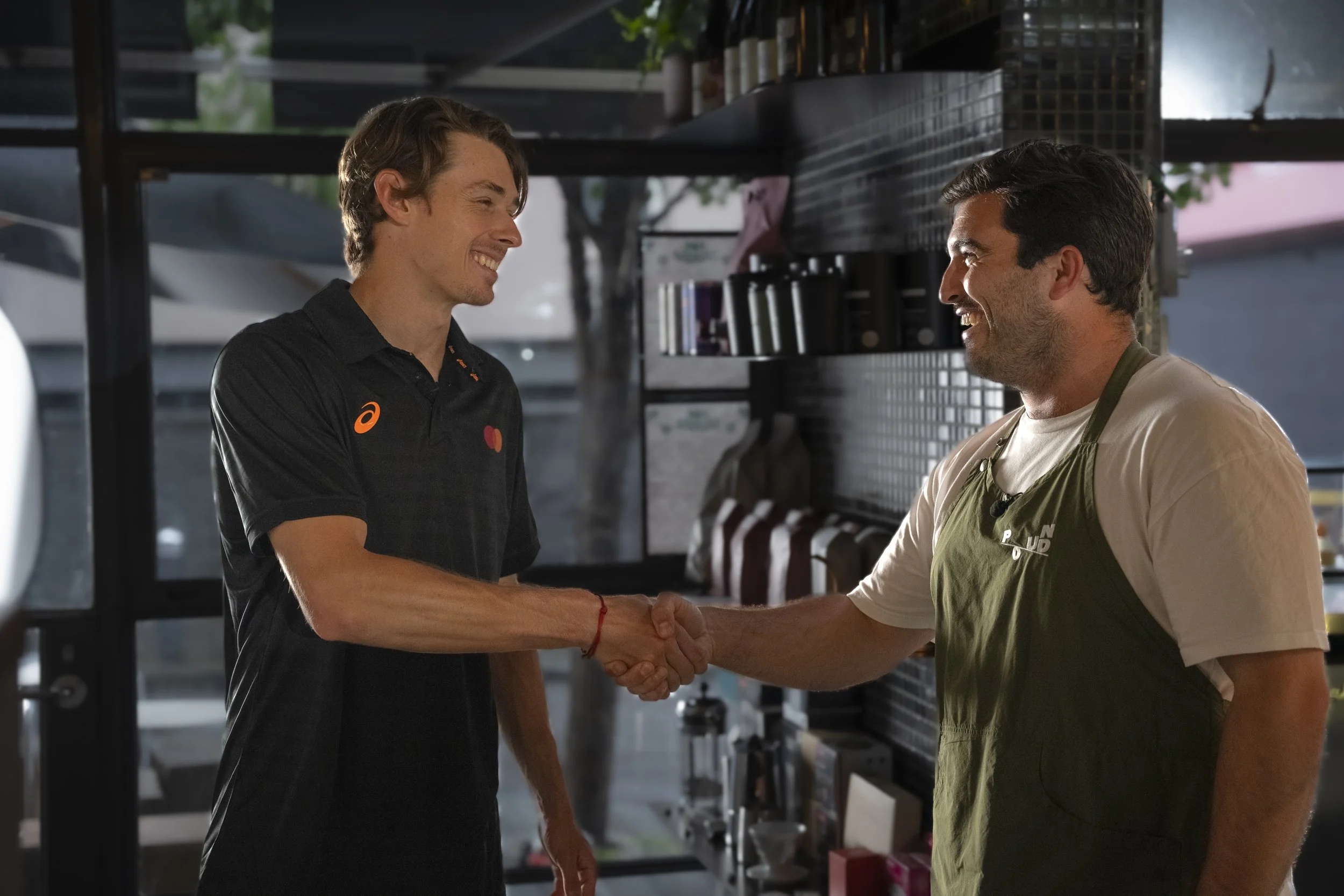 Two men shaking hands and smiling inside a cafe or restaurant, with shelves of coffee products behind them.