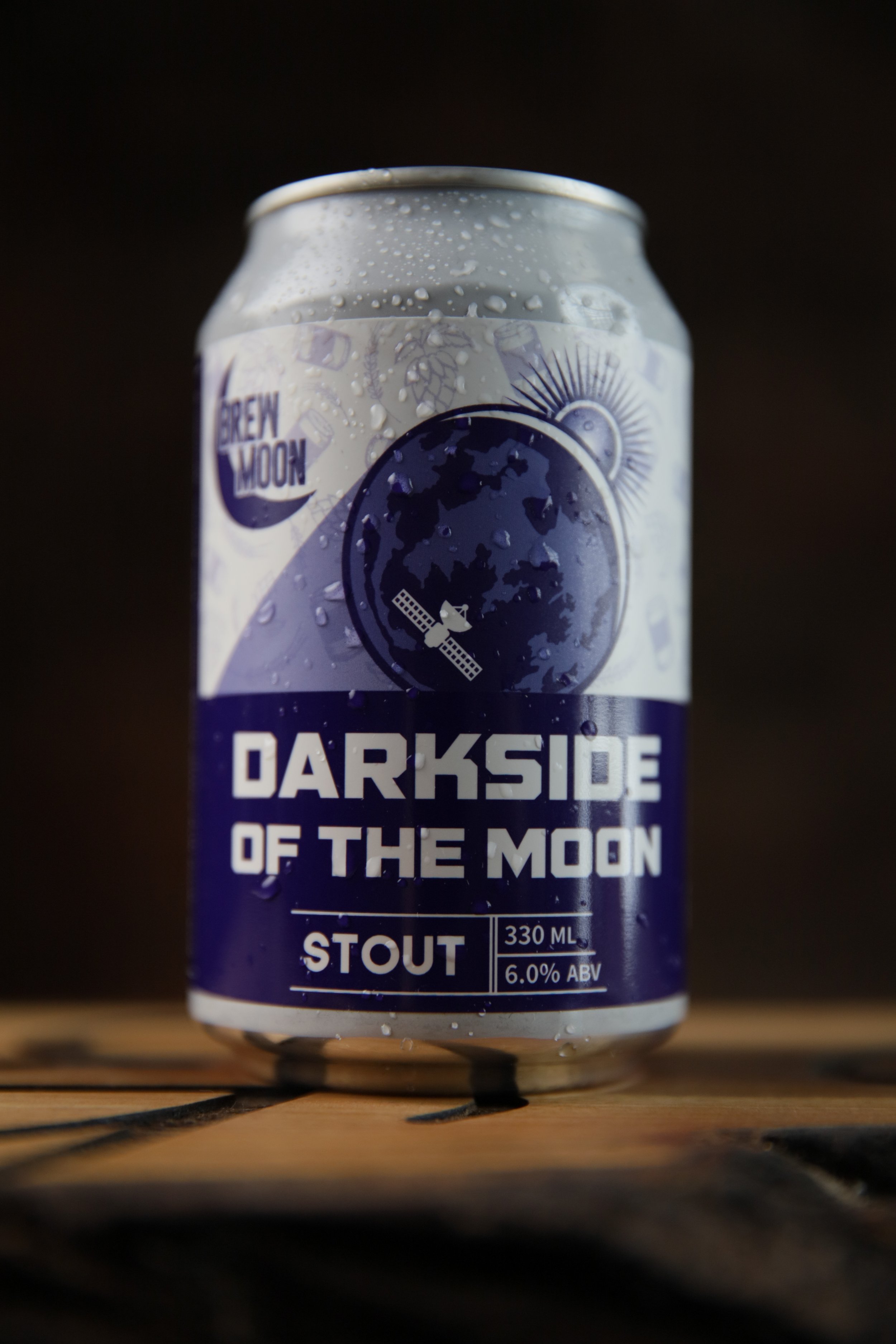 A can of Brew Moon Darkside of the Moon stout beer on a wooden surface with droplets of condensation, featuring a moon and satellite illustration on the label.