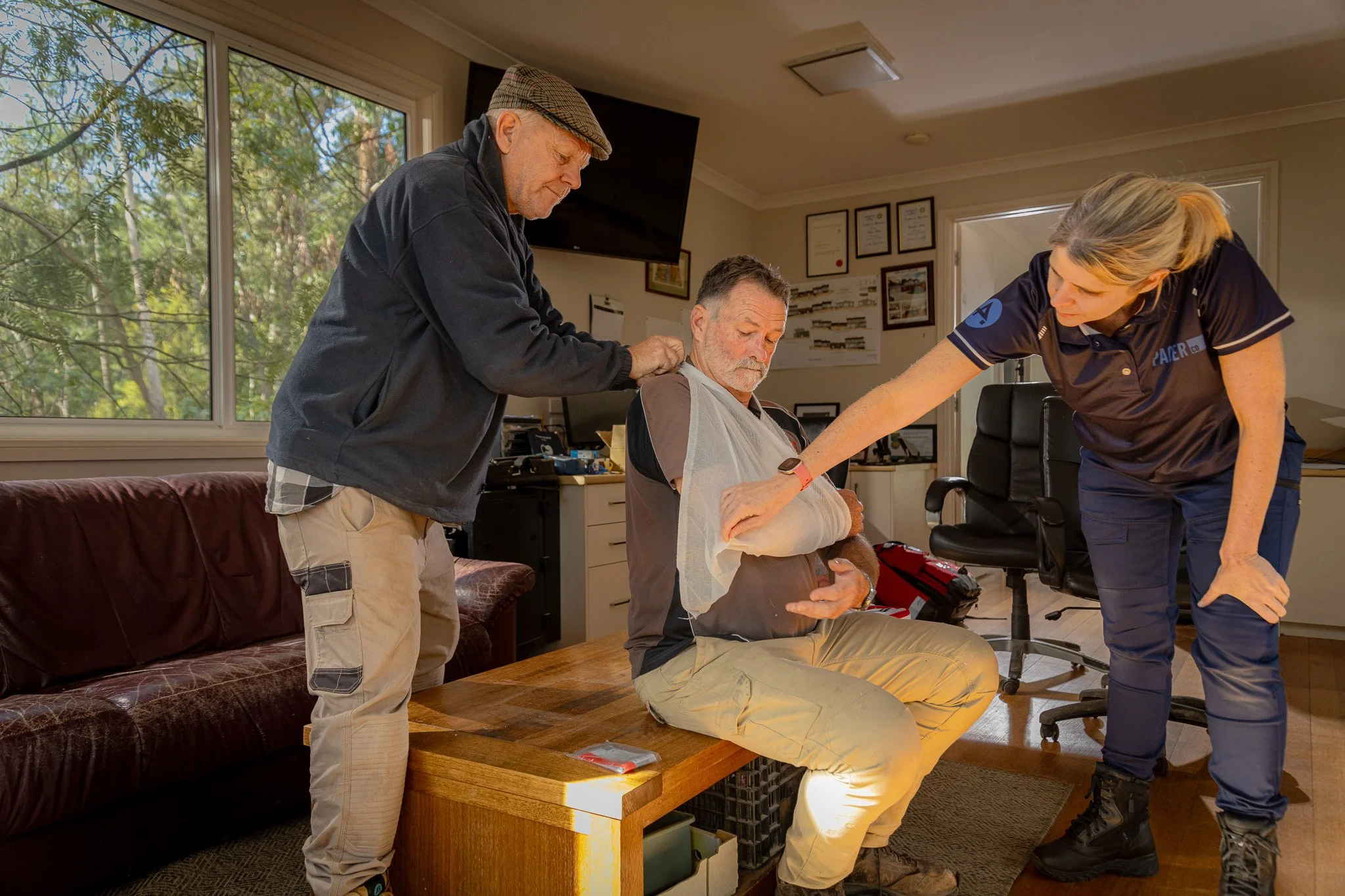 A man sitting on a wooden coffee table with his arm splinted by a woman in a navy uniform and an older man adjusting a medical bandage on his shoulder inside an office or medical facility.