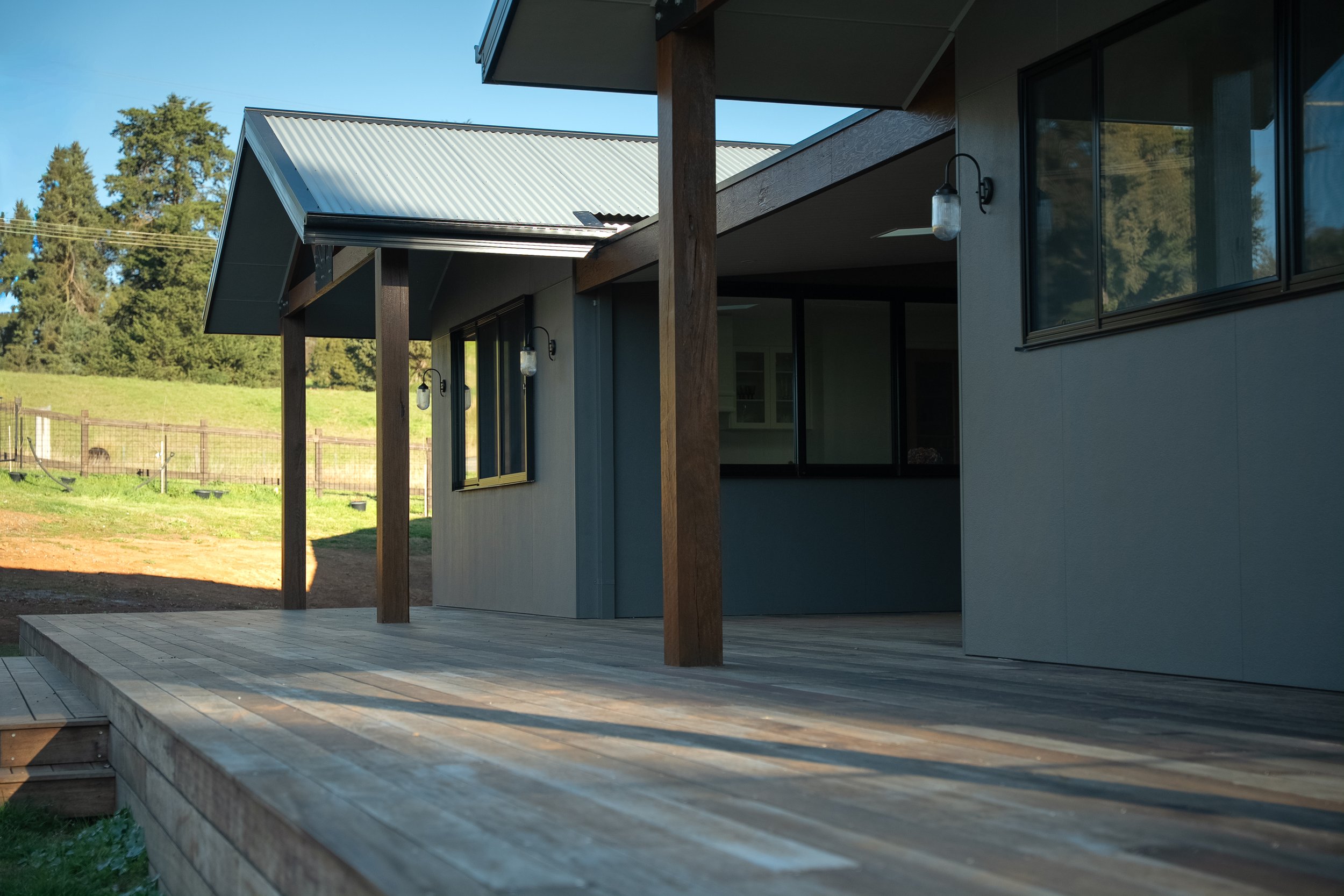 Modern house with a wooden porch, large windows, outdoor lighting, metal roof, surrounded by greenery and a fenced yard.