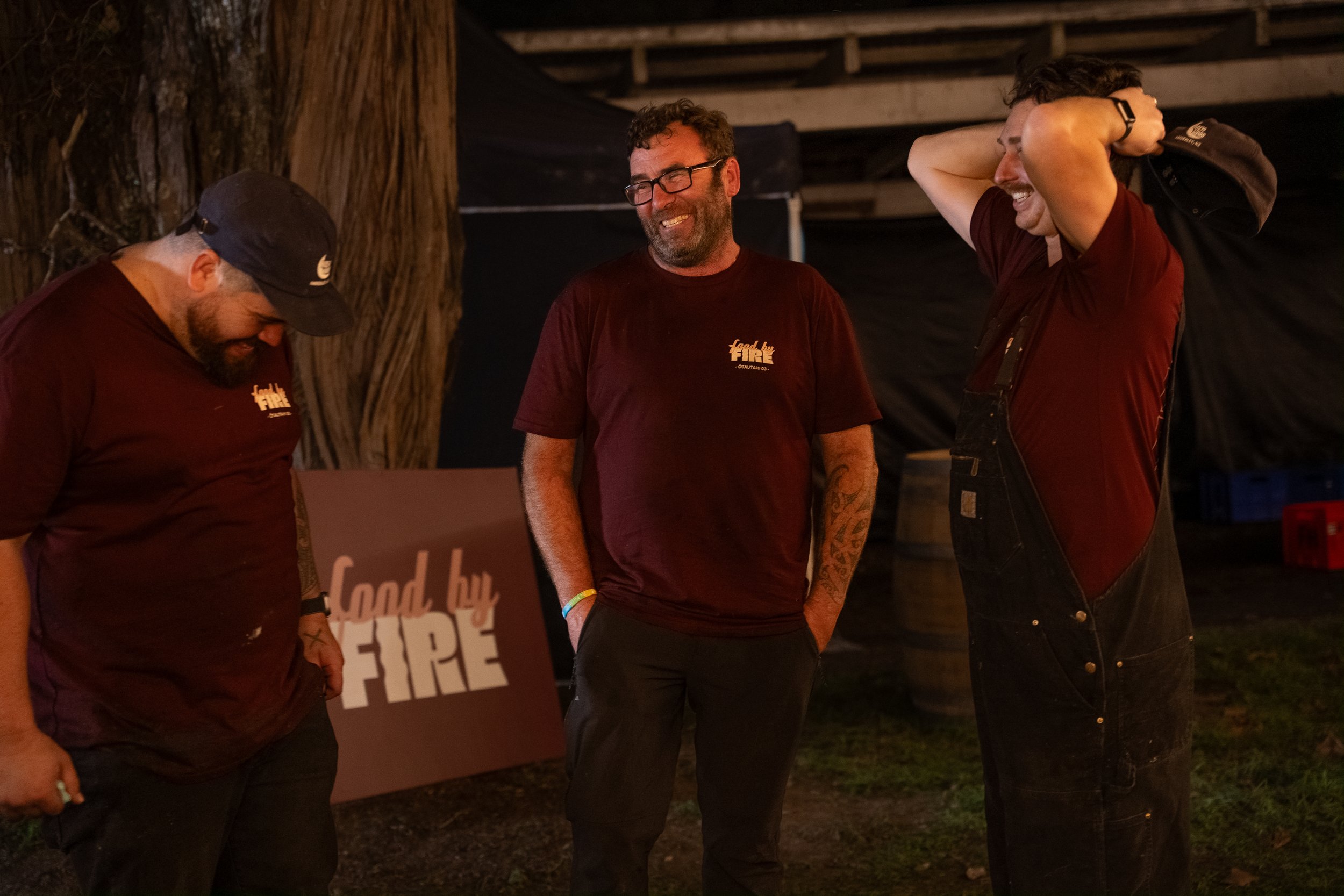 Three men in maroon shirts standing and smiling, in a dimly lit outdoor setting, with a sign that reads 'food by fire' in the background.