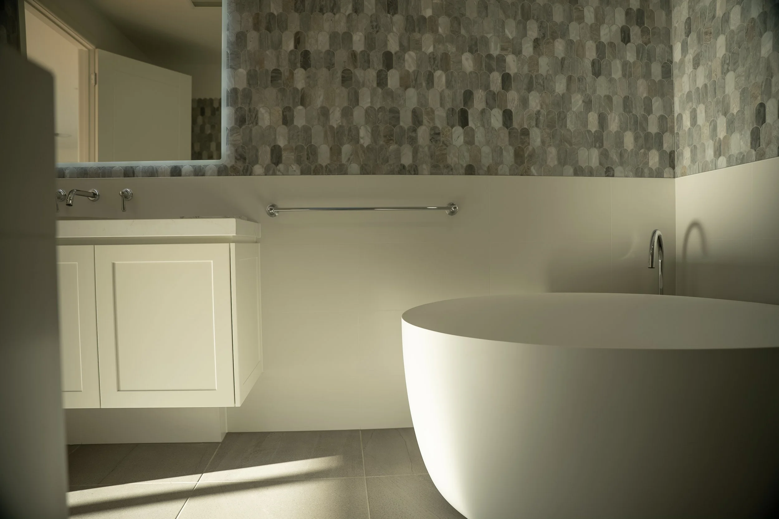 Modern bathroom with a white bathtub, a vanity with a reflective mirror, and a textured wall with gray tiles.