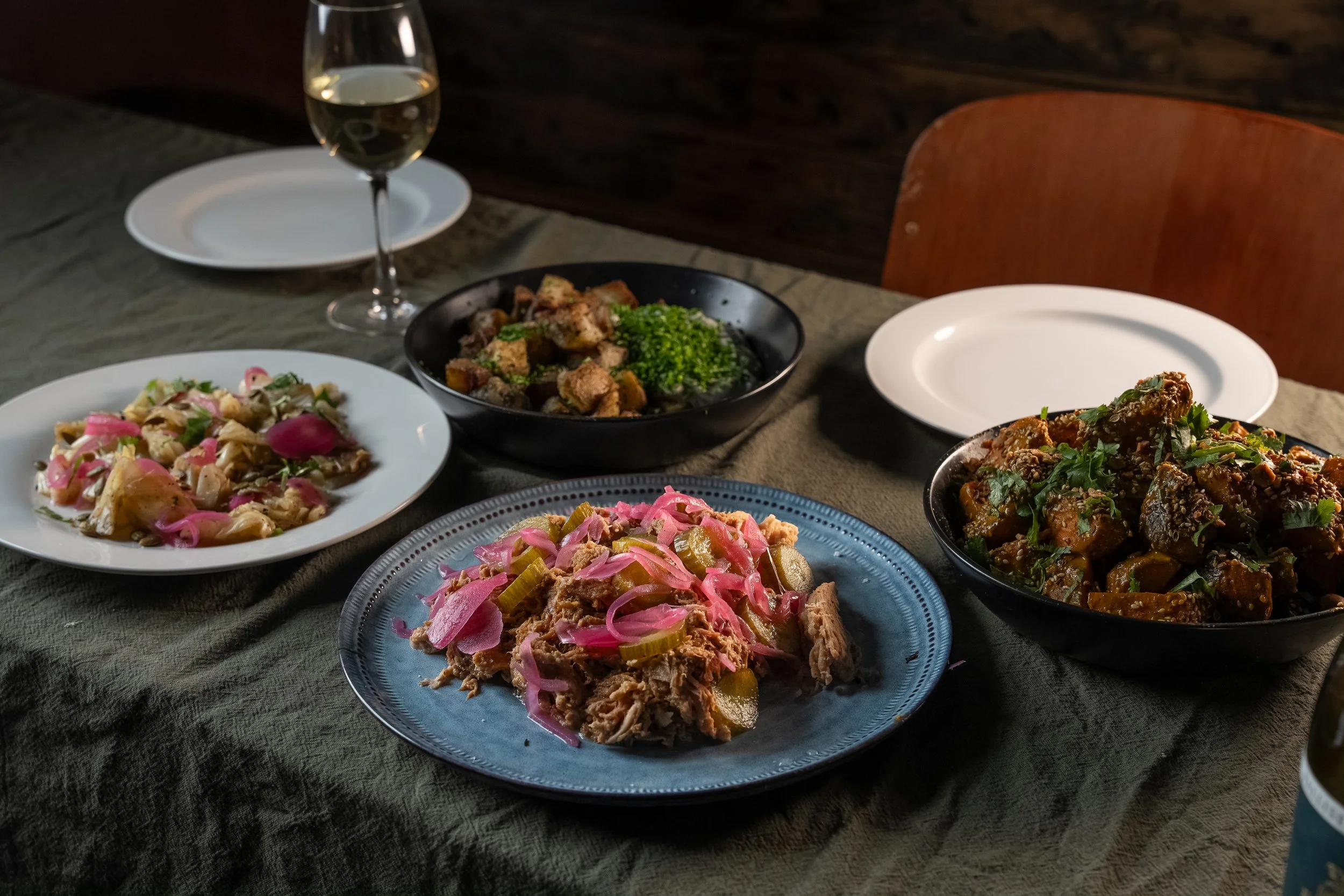 A table with four plates of food, a glass of white wine, and empty plates, in a dimly lit room.