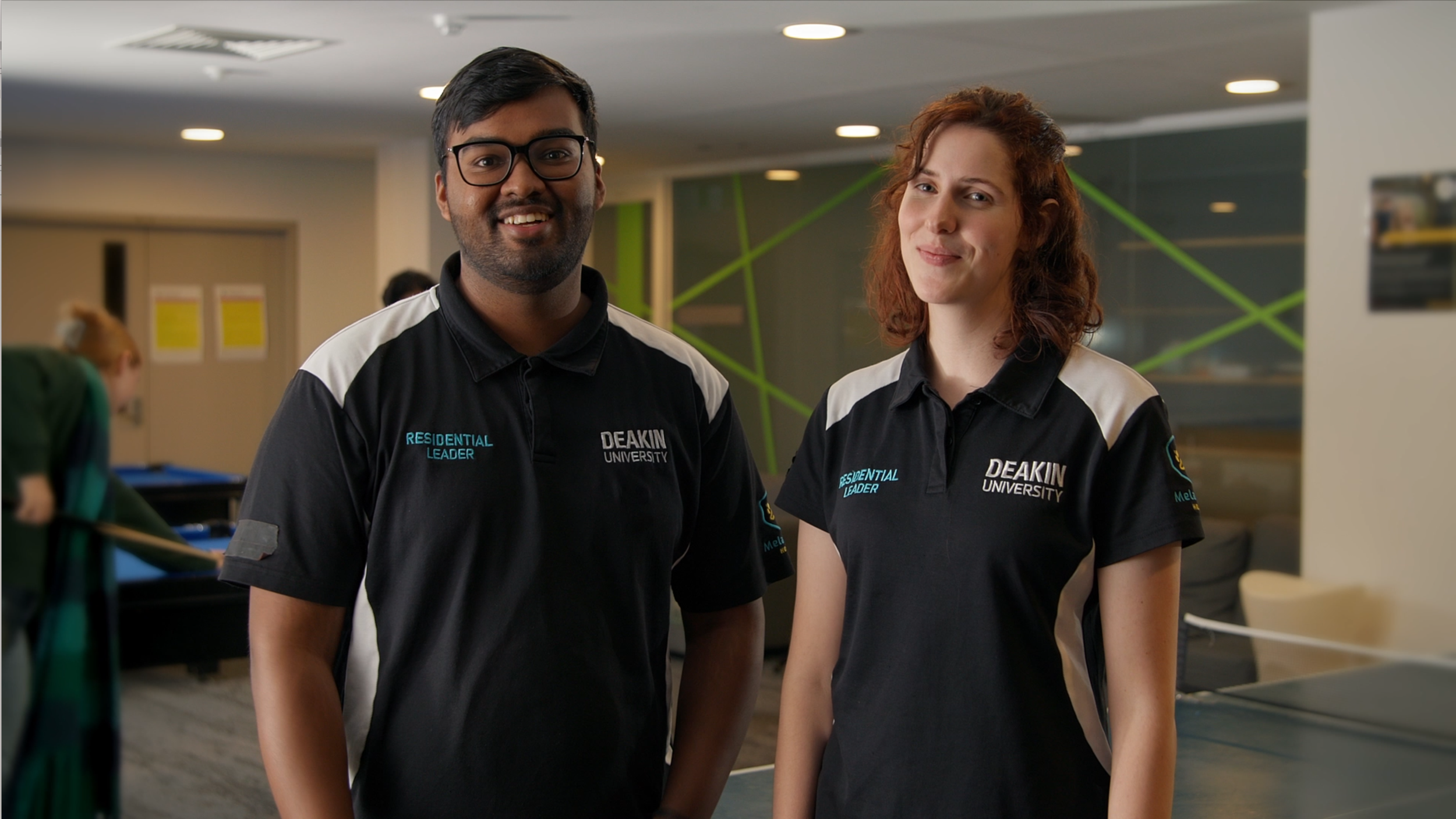 Two students wearing black polo shirts with Deakin University logo pose for a photo in a student lounge, with a ping pong table and other students in the background.