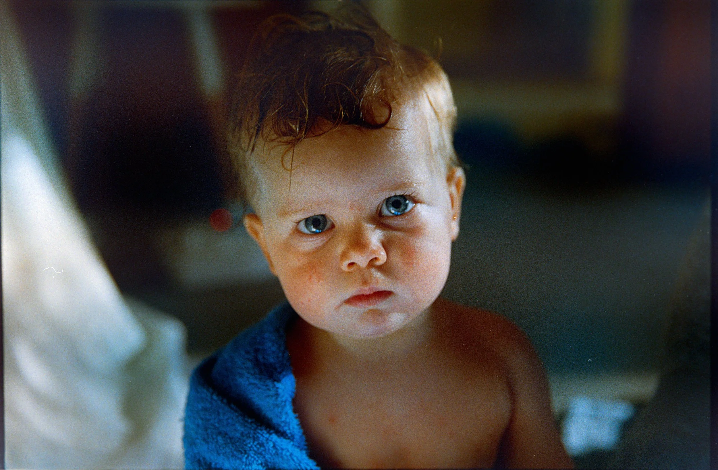 Close-up of a young child with blue eyes, damp brown hair, and a serious expression, wrapped in a blue towel.